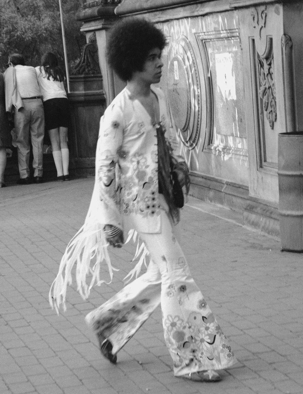 Indivíduo com cabelo afro usa roupa estampada, incluindo calças boca de sino. Ambiente urbano anos 70.