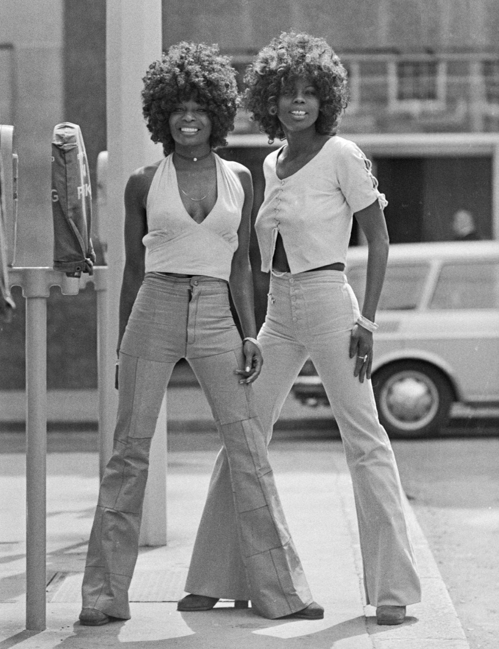 Duas pessoas posam sorrindo, ambas usando calças jeans de cintura alta e estilo anos 70.