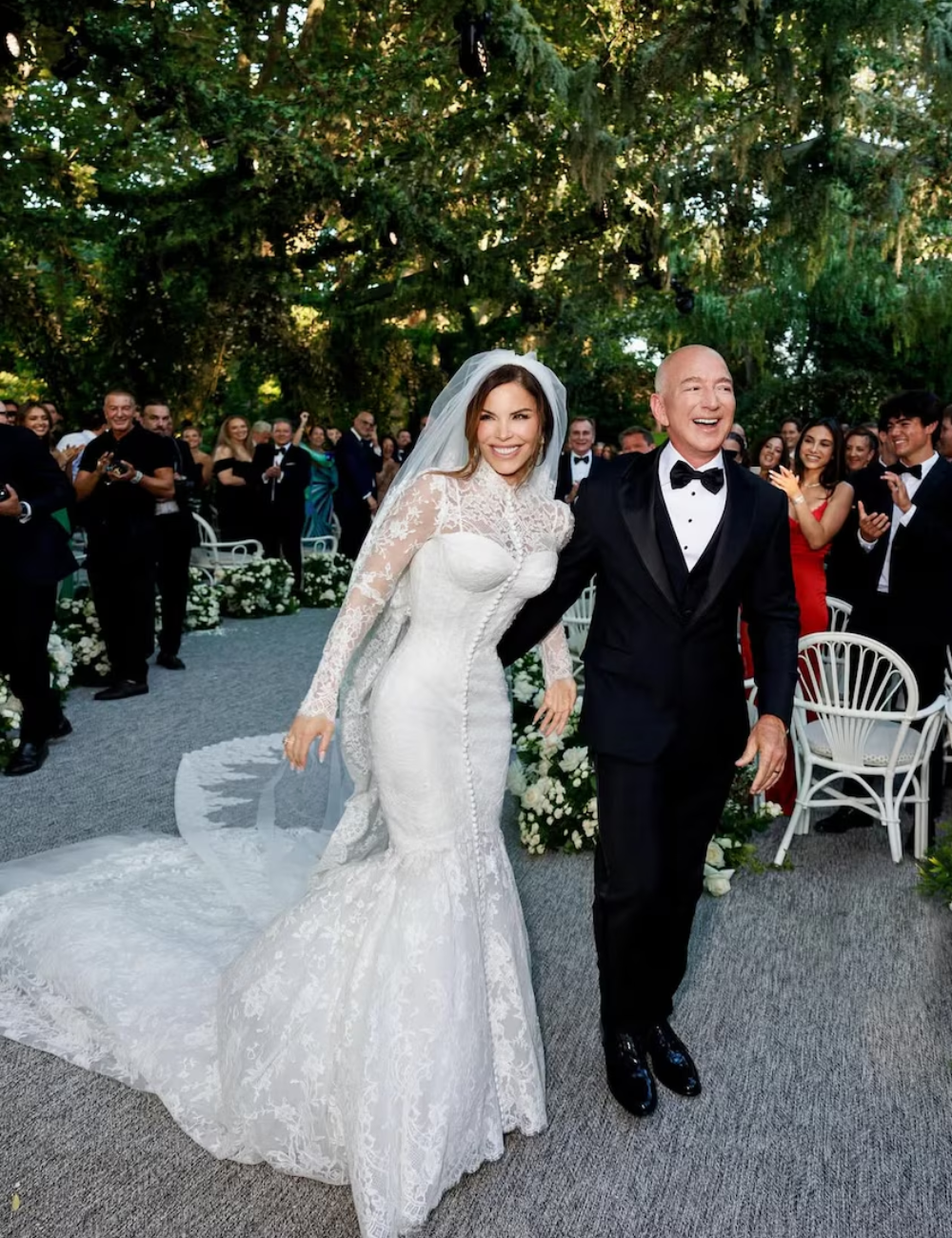 Noiva sorridente, vestida de branco rendado, ao lado de um noivo em terno, em cerimônia ao ar livre.