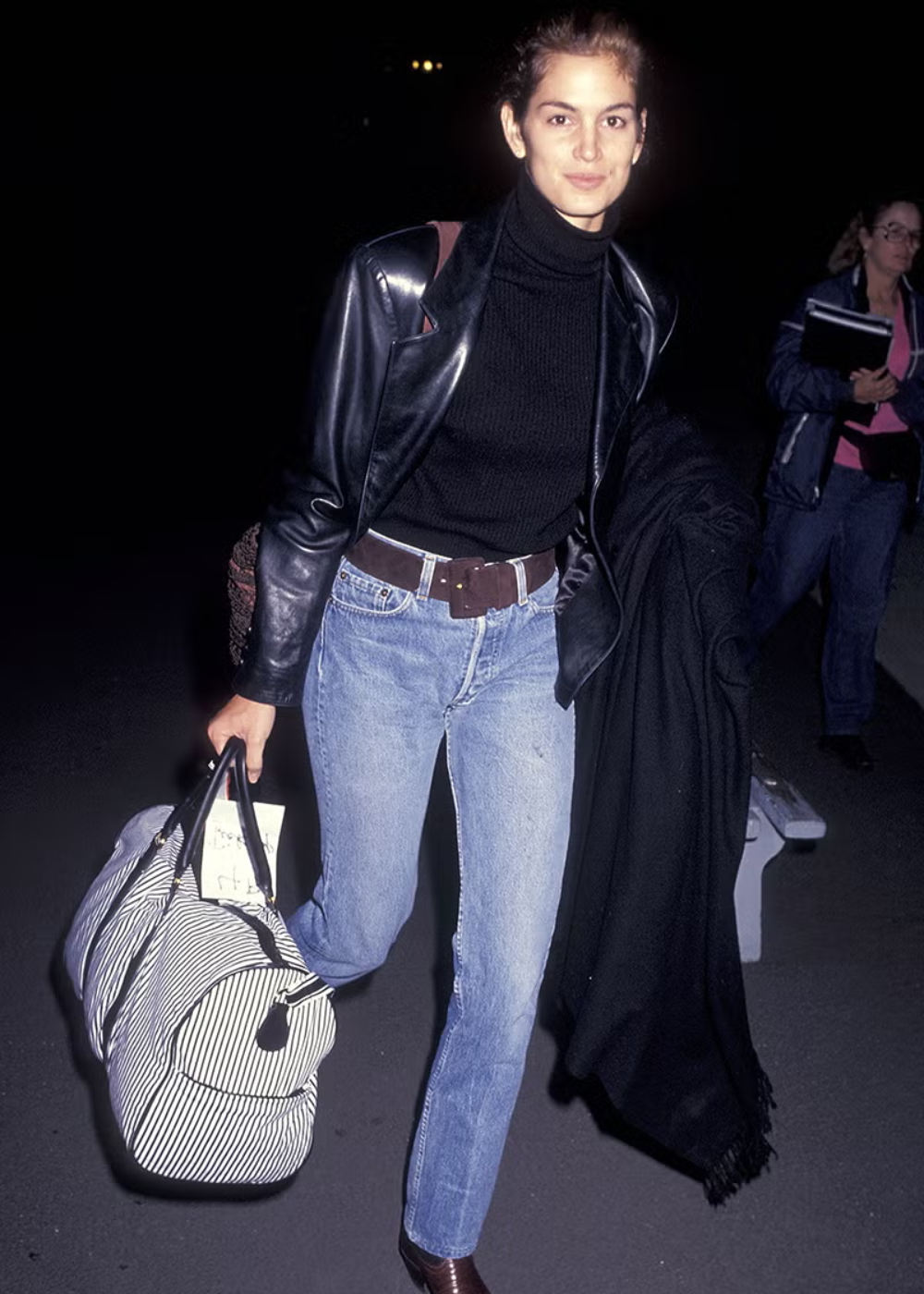 Pessoa usando jaqueta preta e calças jeans tendência, carregando bolsa listrada, em uma rua à noite.