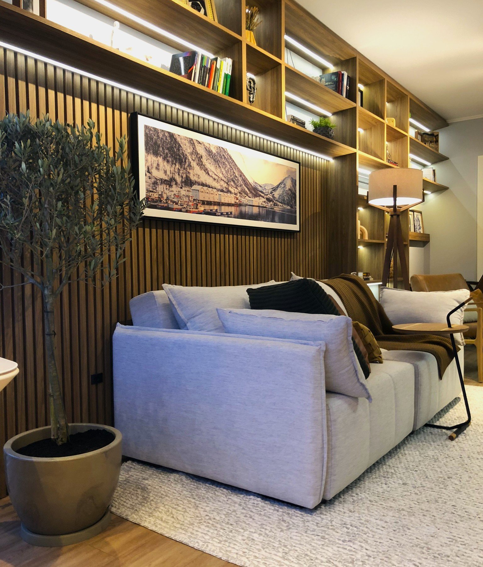 Sala com sofá cinza, estante de madeira, luminária de chão e planta em vaso, criando um ambiente de casa aconchegante.