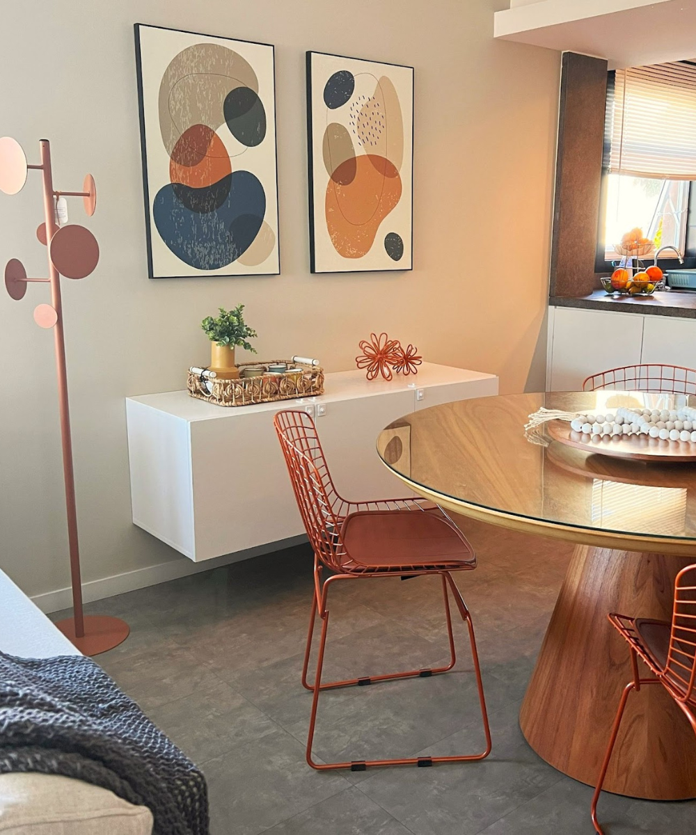 Sala de jantar moderna com mesa de madeira e cadeiras de arame laranja. Quadros abstratos decoram a parede. Casa aconchegante.