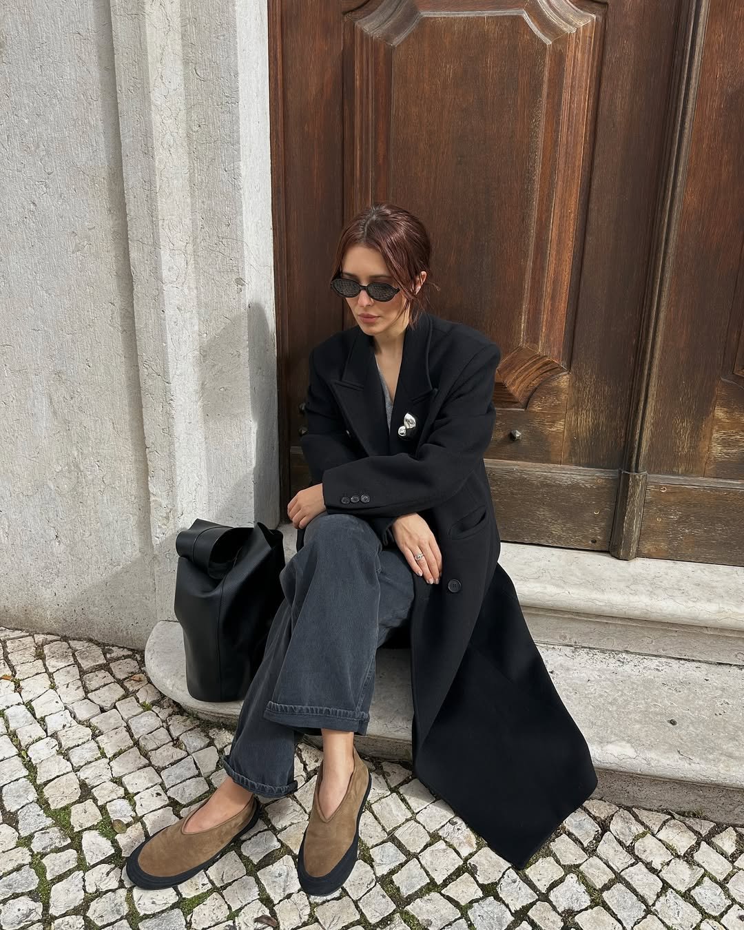 Pessoa sentada em frente a uma porta de madeira, usando casaco feminino de lã batida e óculos escuros, com bolsa ao lado.