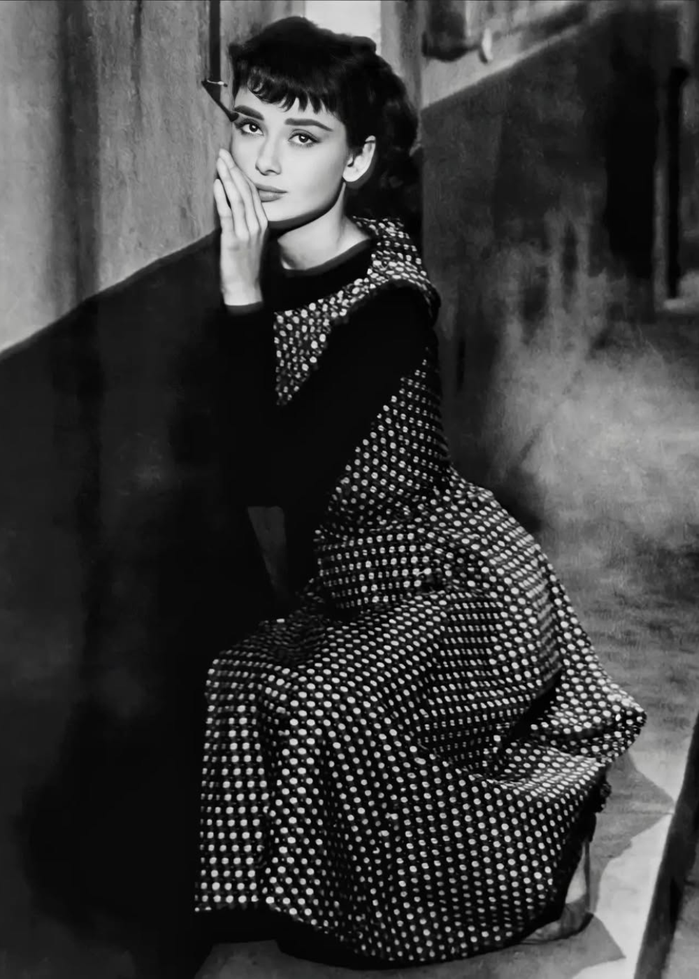 Mulher sentada em um corredor, vestindo um vestido de bolinha, olhando pensativa para a frente.