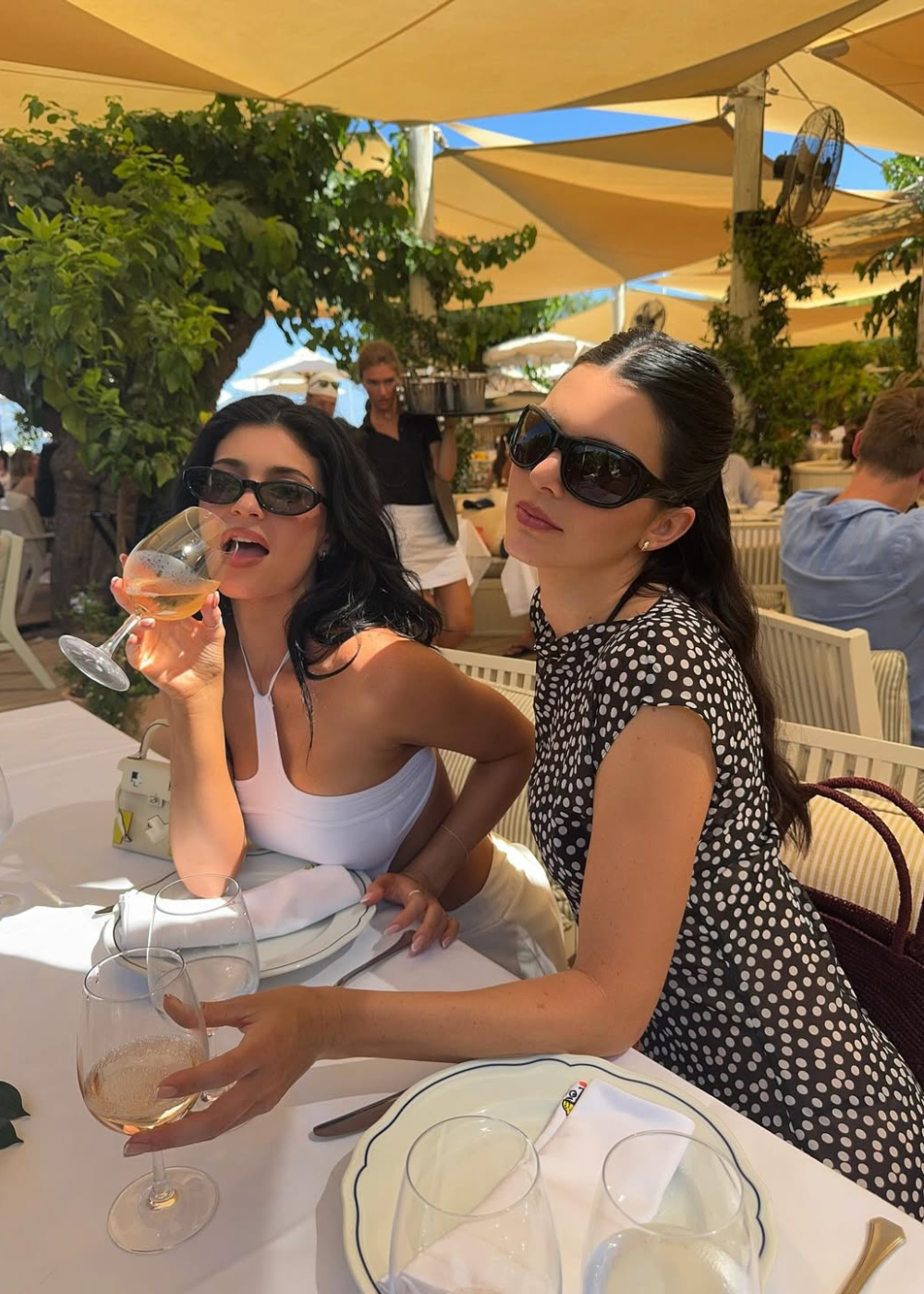 Duas mulheres em um restaurante ao ar livre; uma usa vestido de bolinha e óculos escuros.