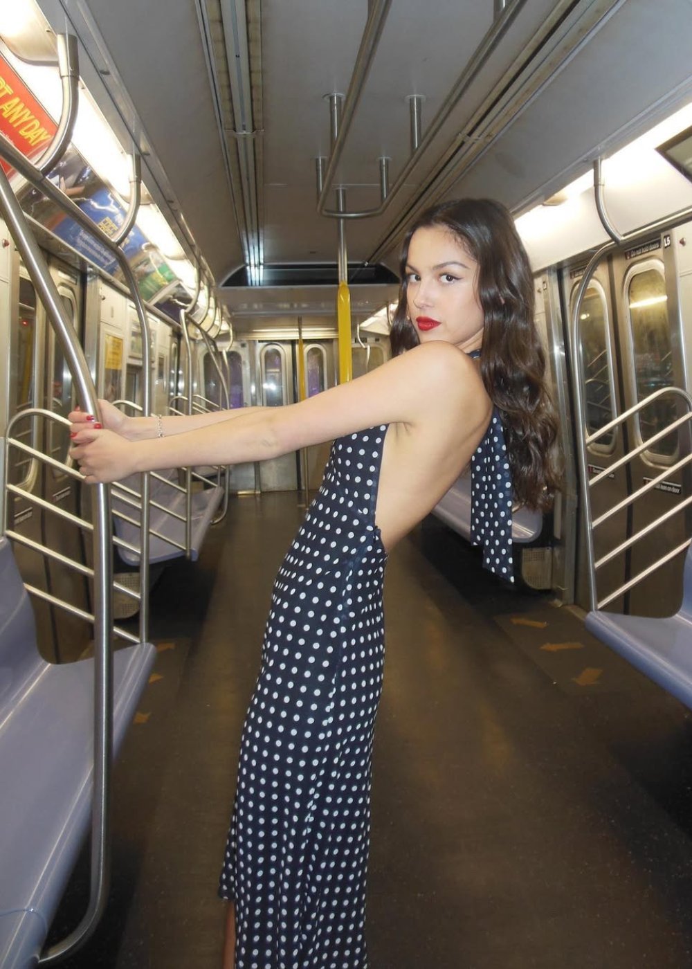 Mulher em um vestido de bolinha preto e branco em um vagão de metrô vazio, segurando uma barra de suporte.