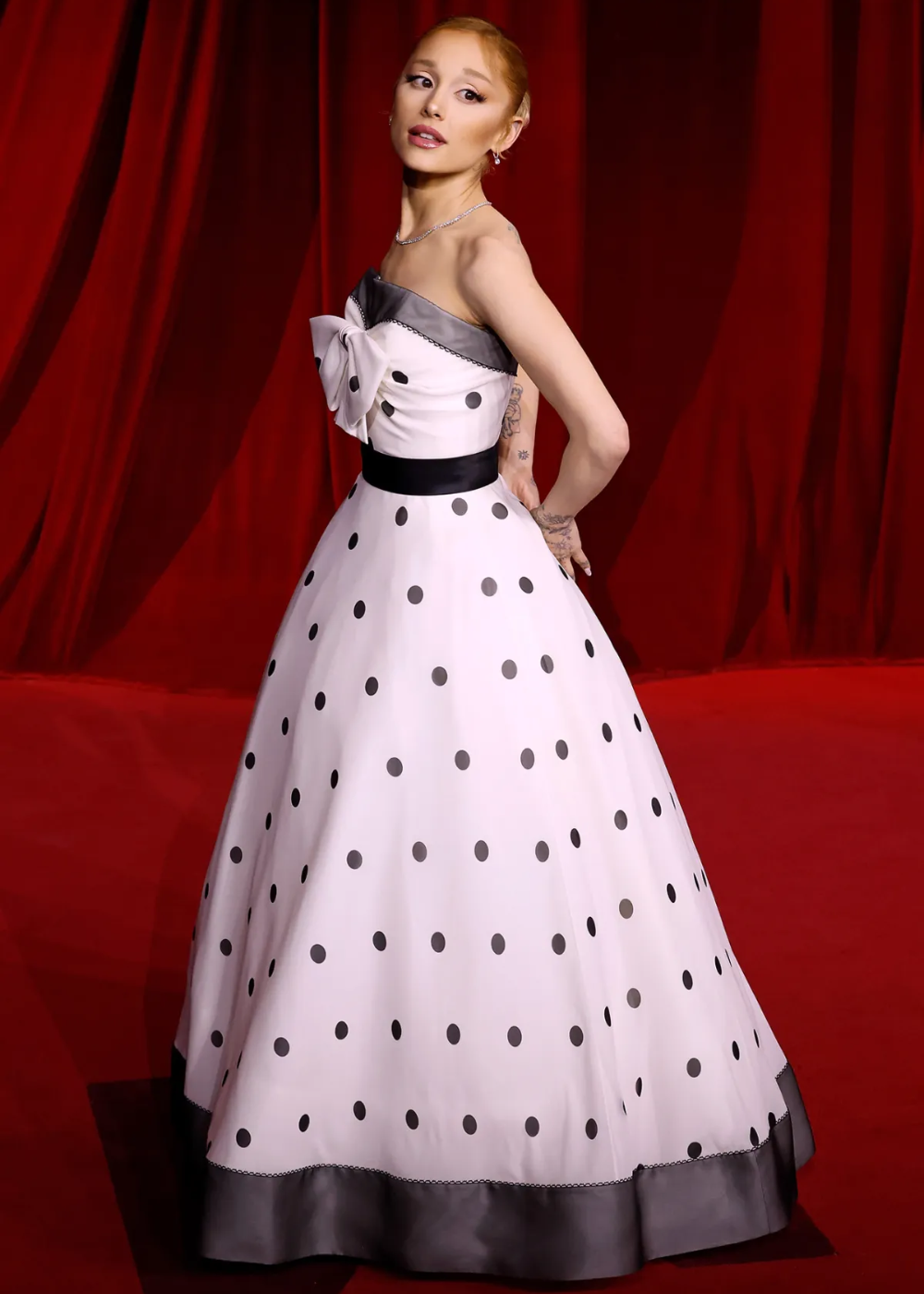 Mulher em vestido de bolinha preto e branco com laço, em frente a uma cortina vermelha.