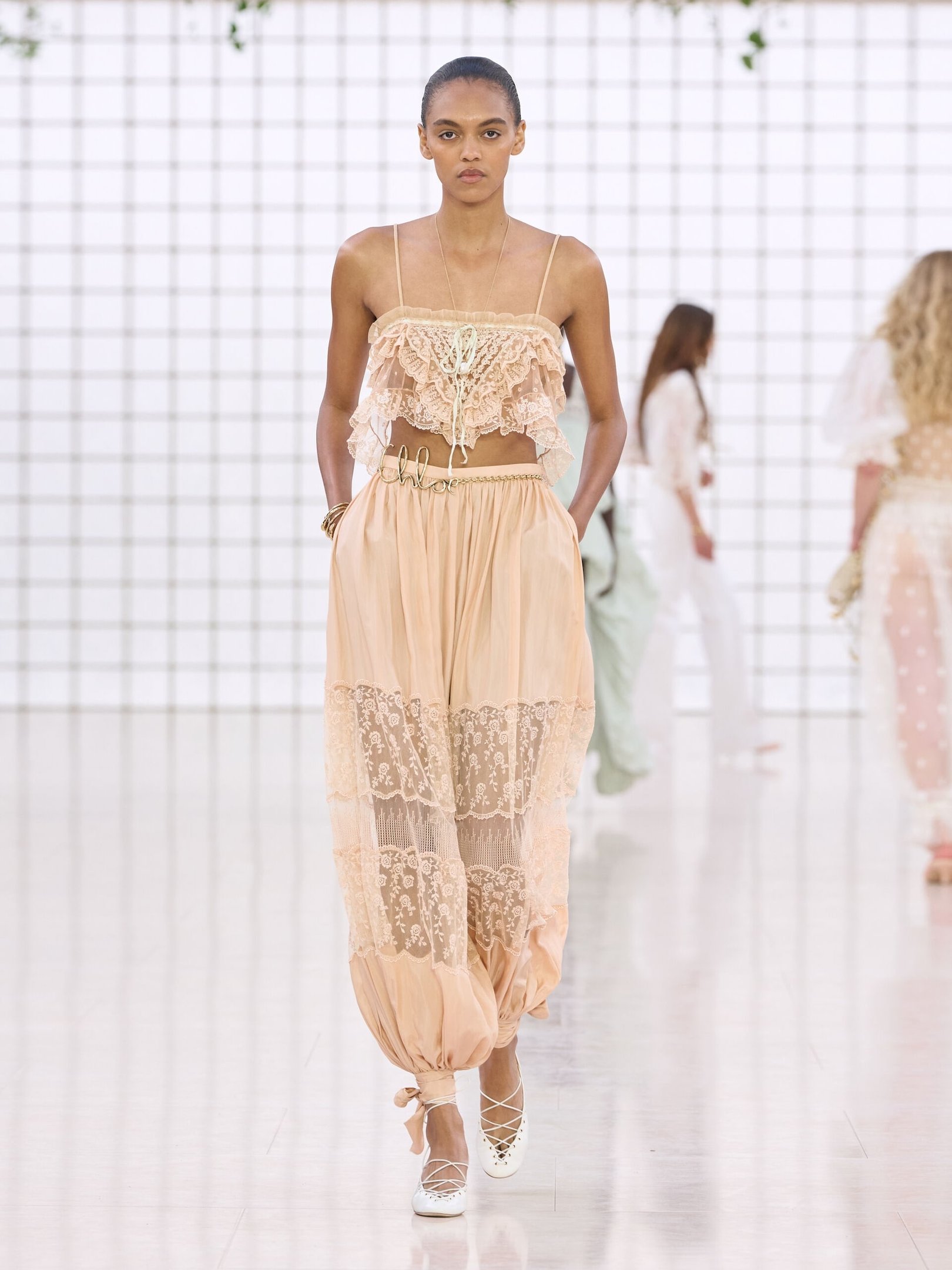 Desfile de moda com modelo vestindo top rendado e calça de estilo balonê em tom bege claro.