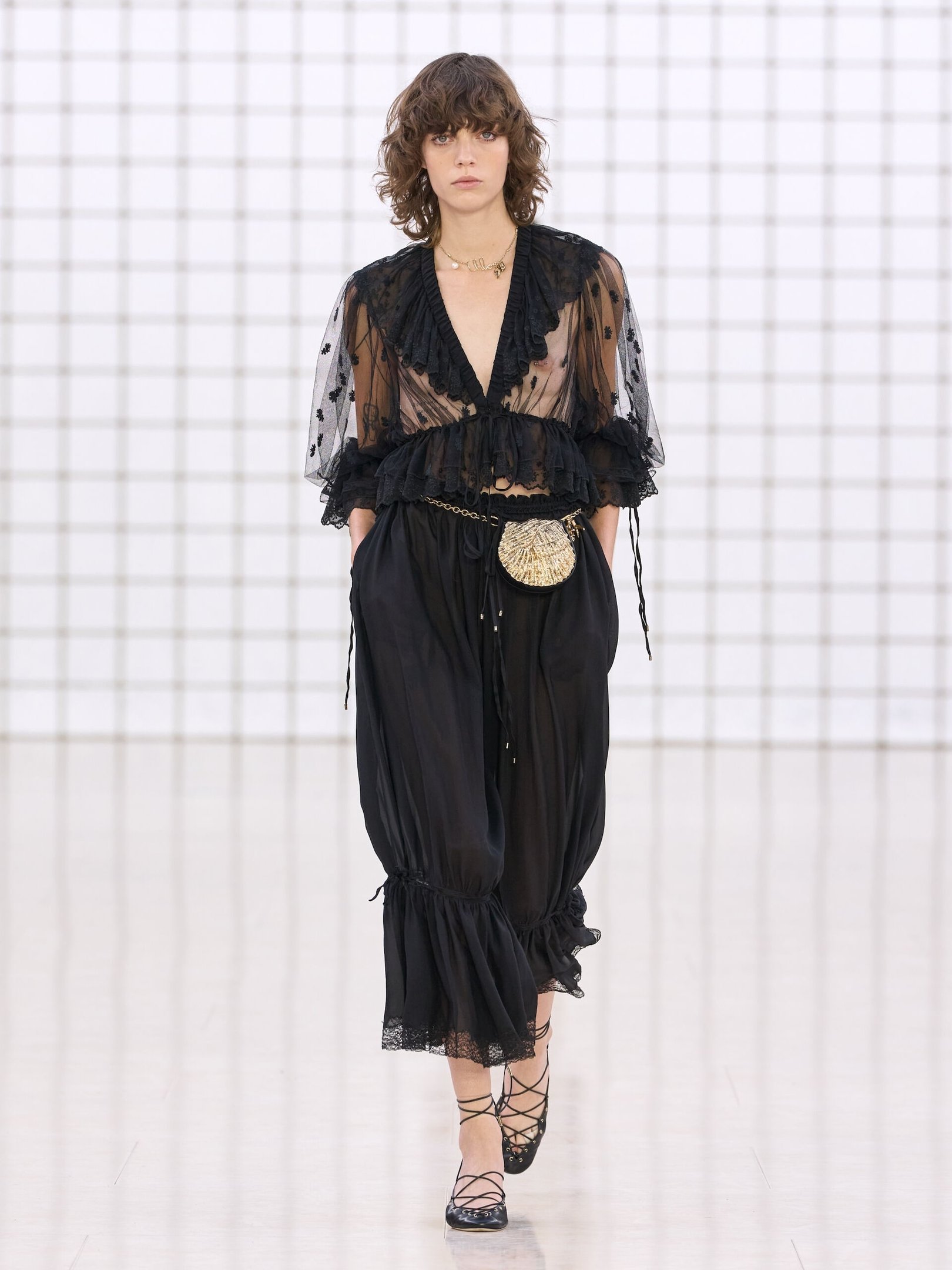 Modelo com blusa transparente e calça balonê preta, usa bolsa pequena e sapatos de amarração.