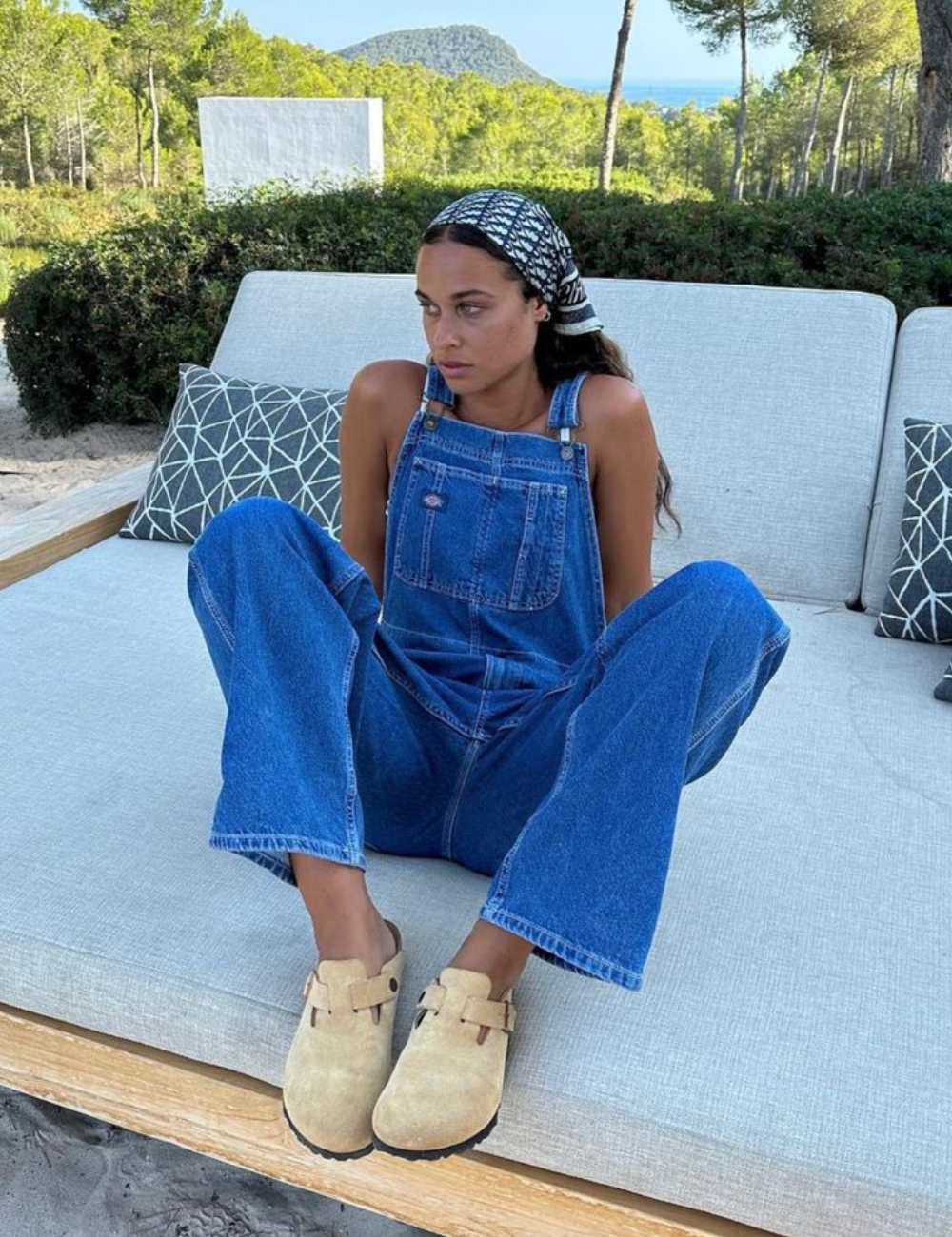 Mulher de macacão jeans e bandana sentada em sofá ao ar livre, vestindo clogs bege.