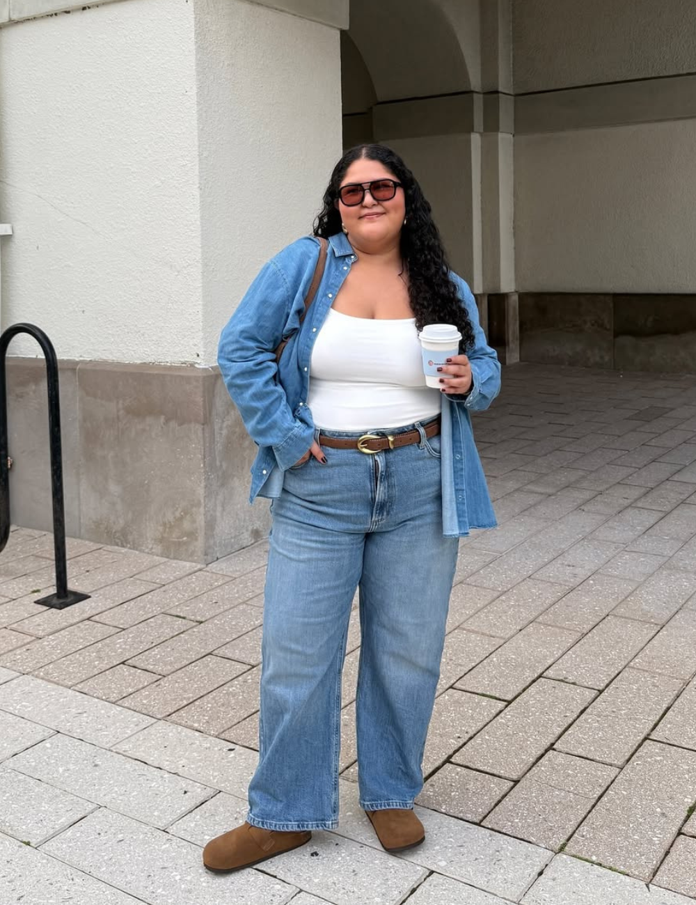 Pessoa de óculos escuros, camisa jeans e calça larga, segurando café. Ela usa clogs marrom em uma calçada.