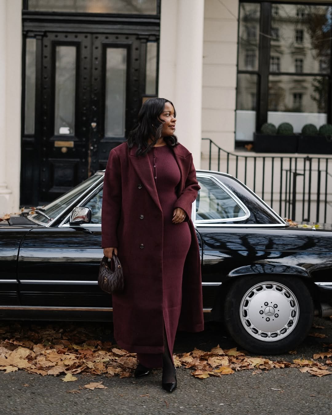 Mulher em casaco e vestido vinho junto a carro preto vintage, rodeada de folhas secas, demonstrando combos de estilo.