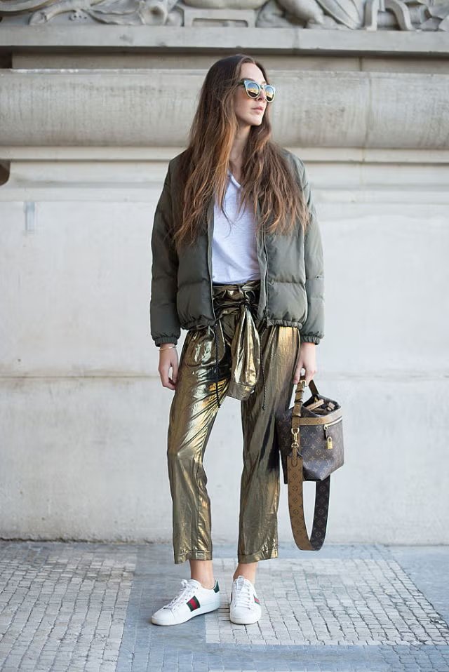 Jovem com jaqueta verde, calça dourada metalizada e tênis brancos, criando combos de estilo ousados. Óculos de sol e bolsa de marca.