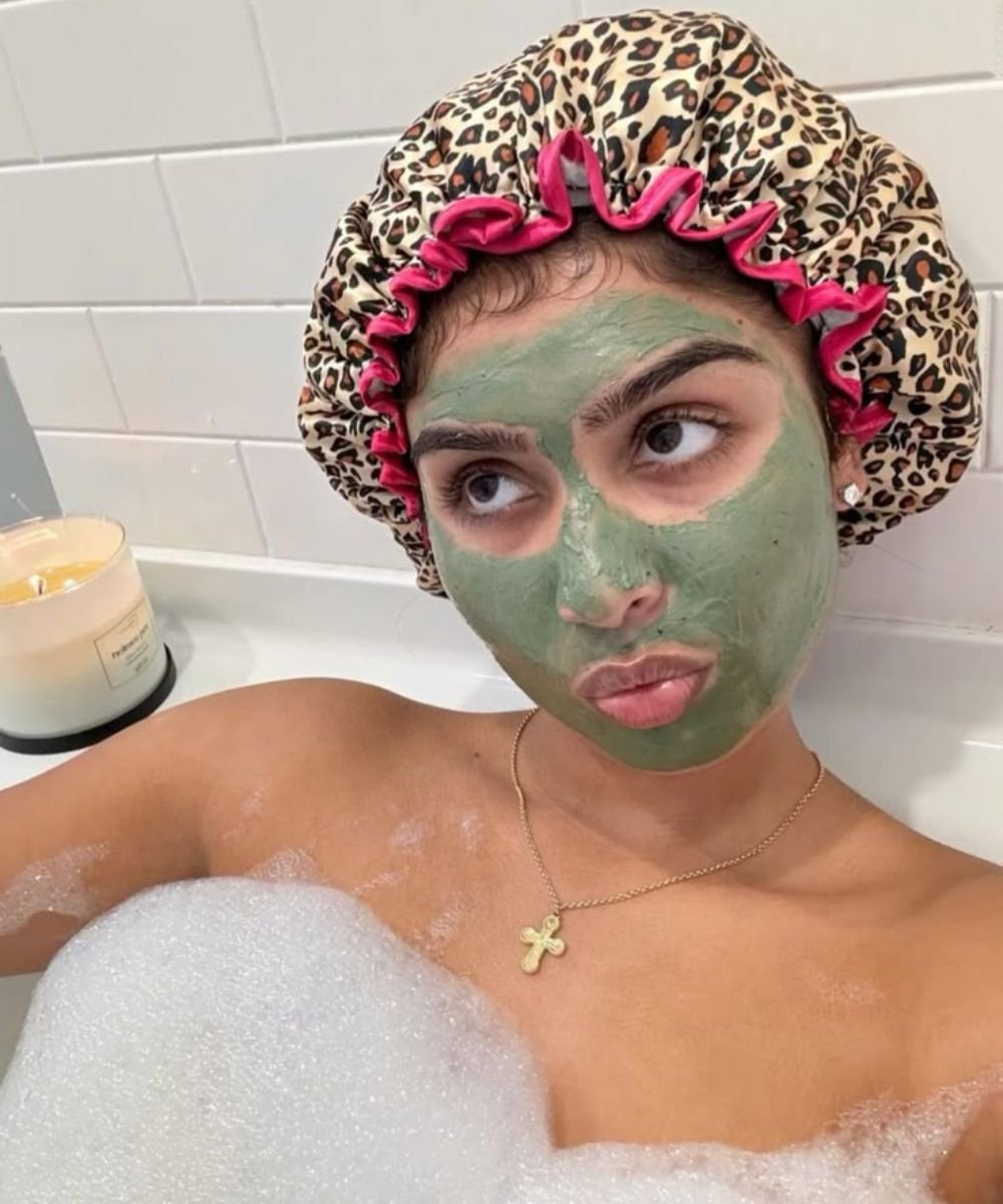 Pessoa relaxando na banheira com espuma, máscara facial verde e touca de oncinha. Vela acesa ao lado. Um day after de autocuidado.