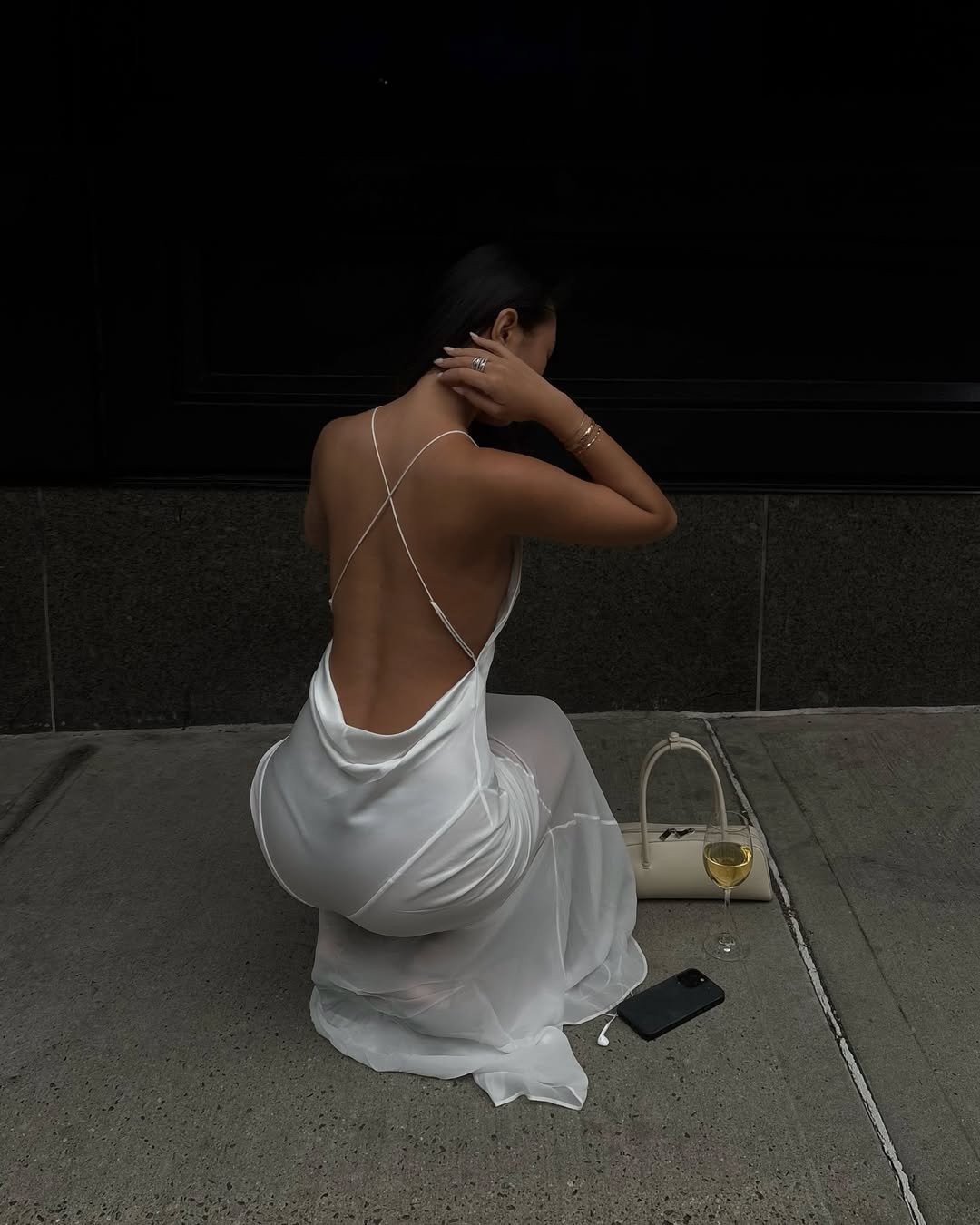 Mulher em vestido branco de alças finas sentada na calçada com bolsa e copo de vinho.