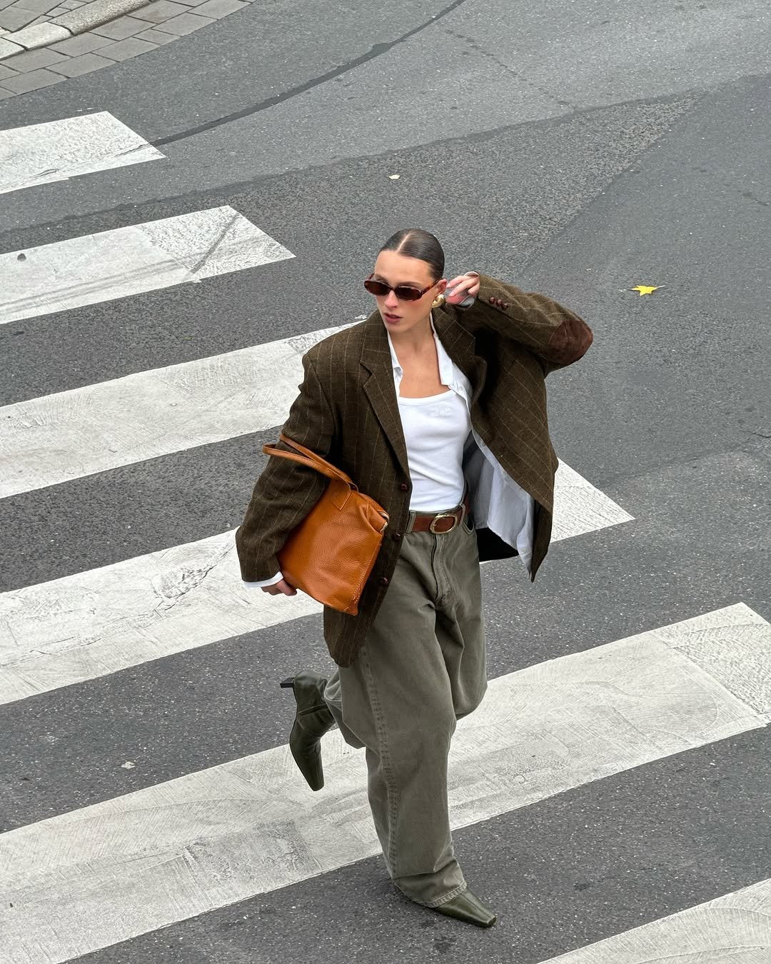 Pessoa com óculos escuros, blazer marrom e bolsa laranja atravessa a rua, sugerindo estilo profissional.