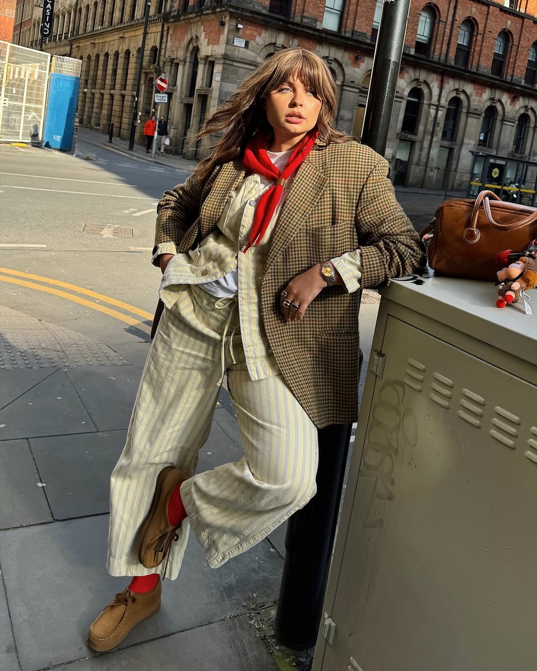 Pessoa de pé na calçada, usando blazer xadrez e calça listrada larga, com expressão pensativa.