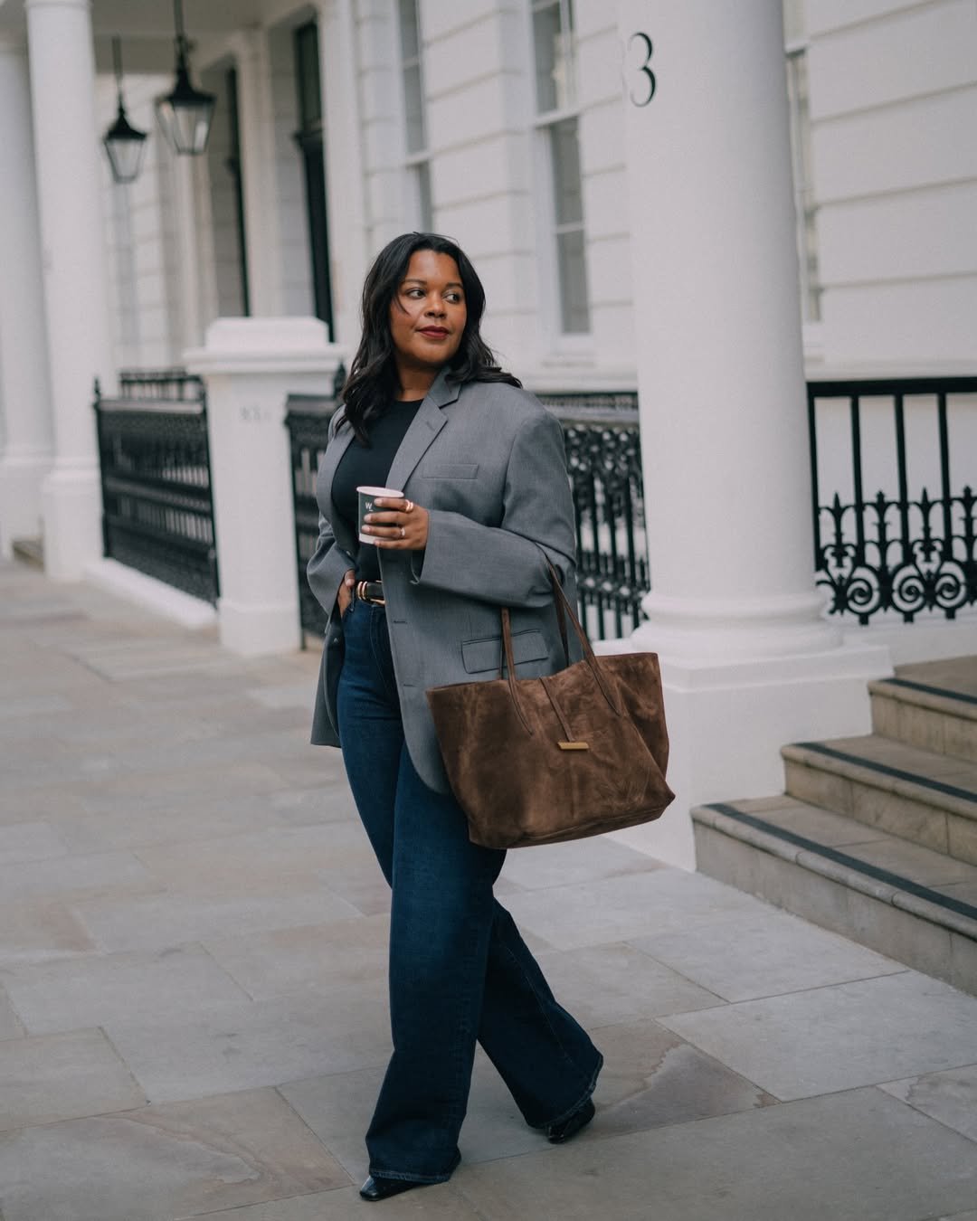 Pessoa andando com blazer cinza e jeans, segurando uma bolsa grande e um café, exemplo de como se vestir para uma entrevista profissional.