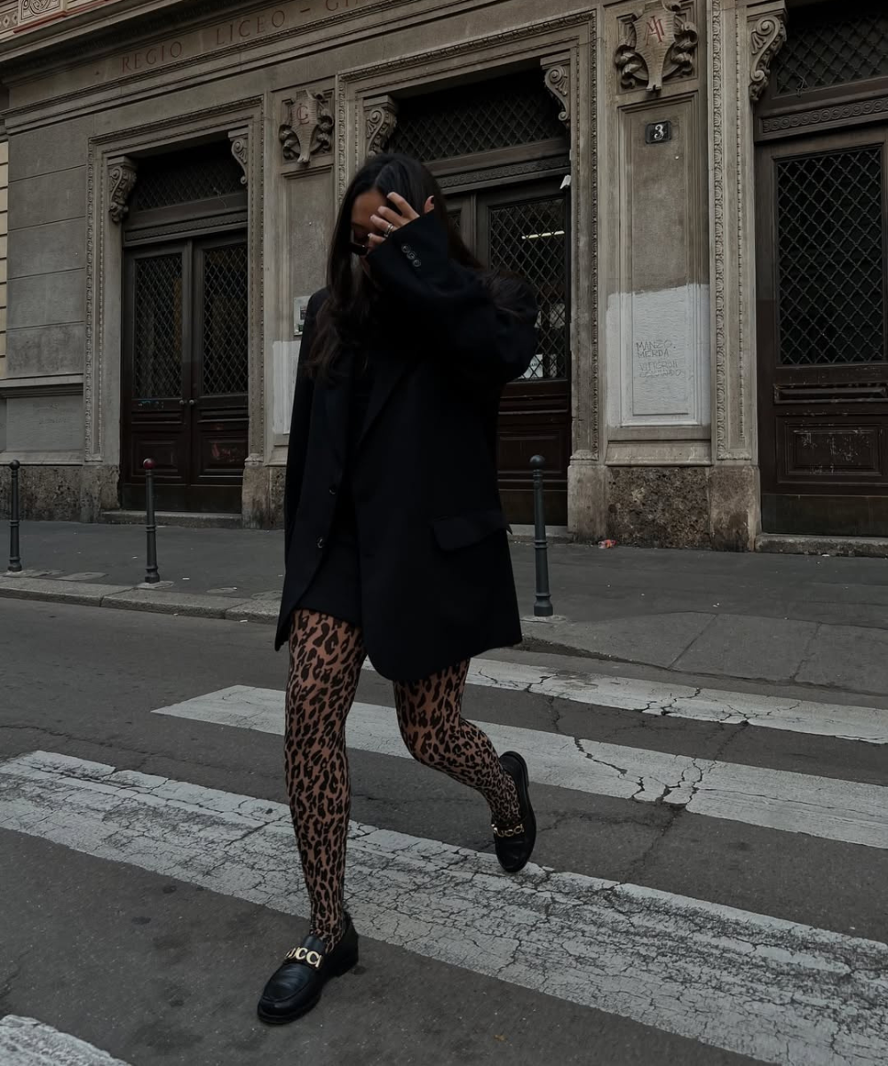 Pessoa andando na rua com casaco preto, meia-calça com estampa de oncinha e mocassins. Fachada de prédio ao fundo.
