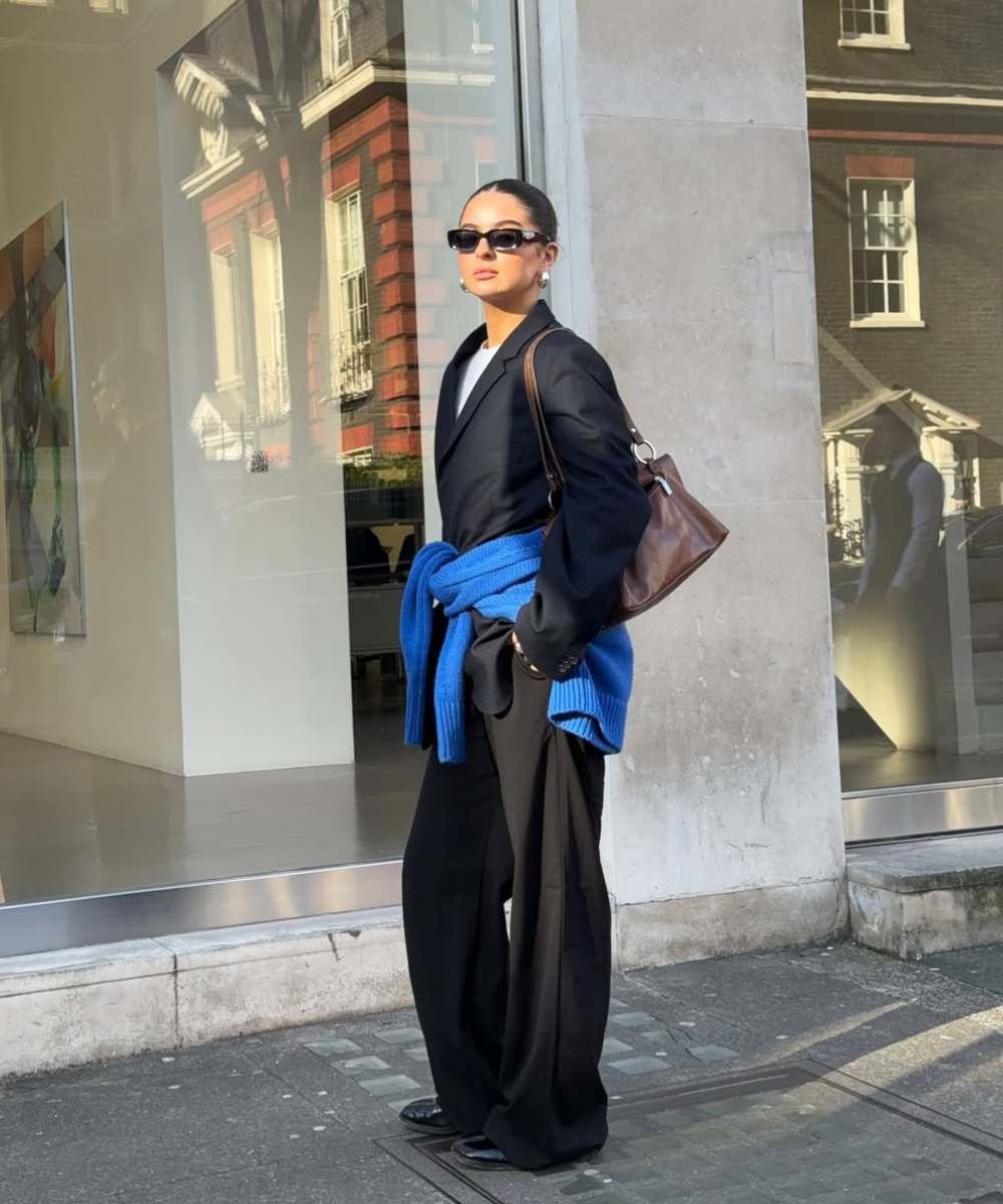 Pessoa usa blazer preto, calças largas, óculos escuros e carrega uma bolsa marrom. Casaco azul amarrado à cintura.