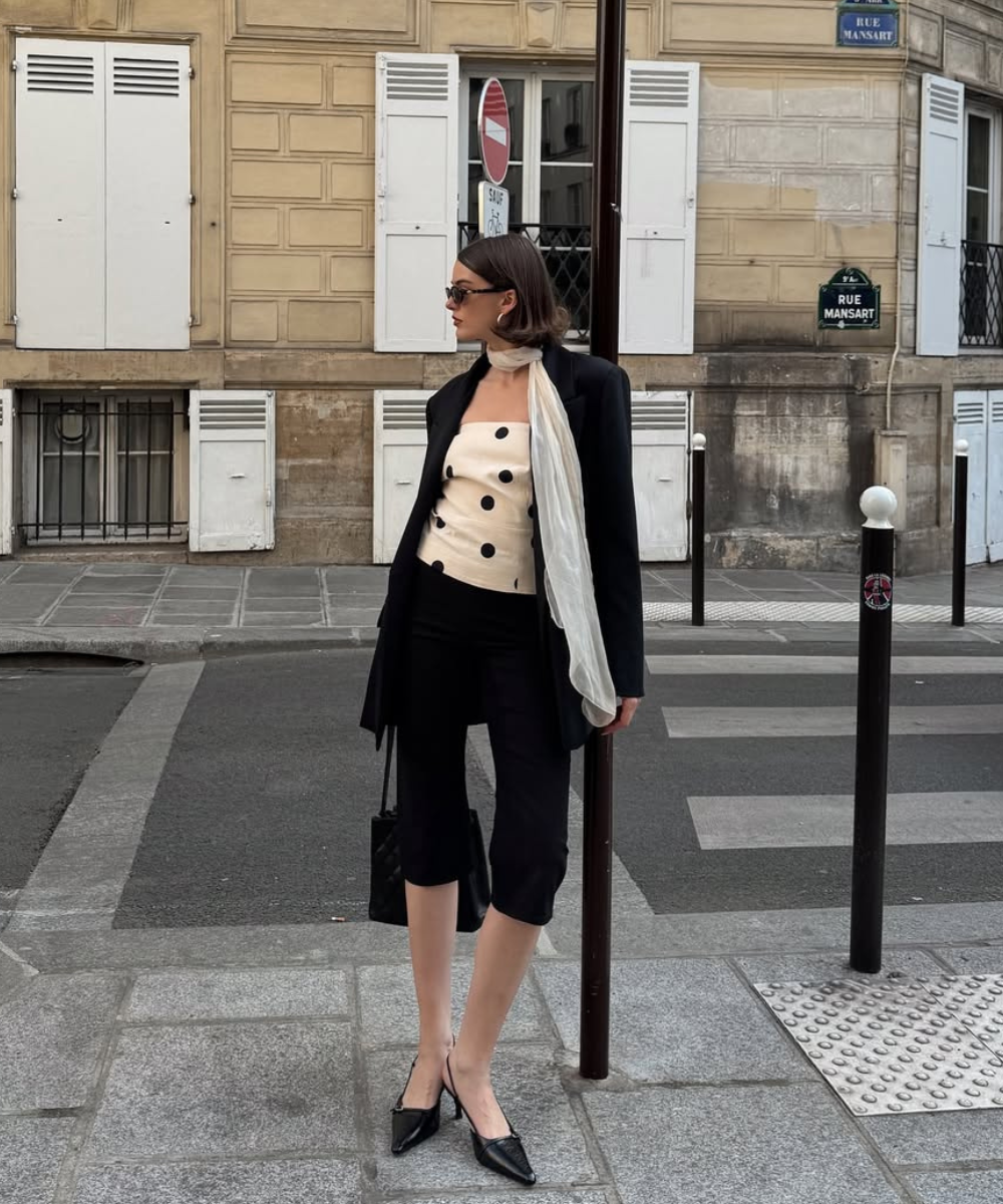 Mulher vestindo blazer preto e calça cropped, segurando bolsa, em rua de cidade com edifícios ao fundo.