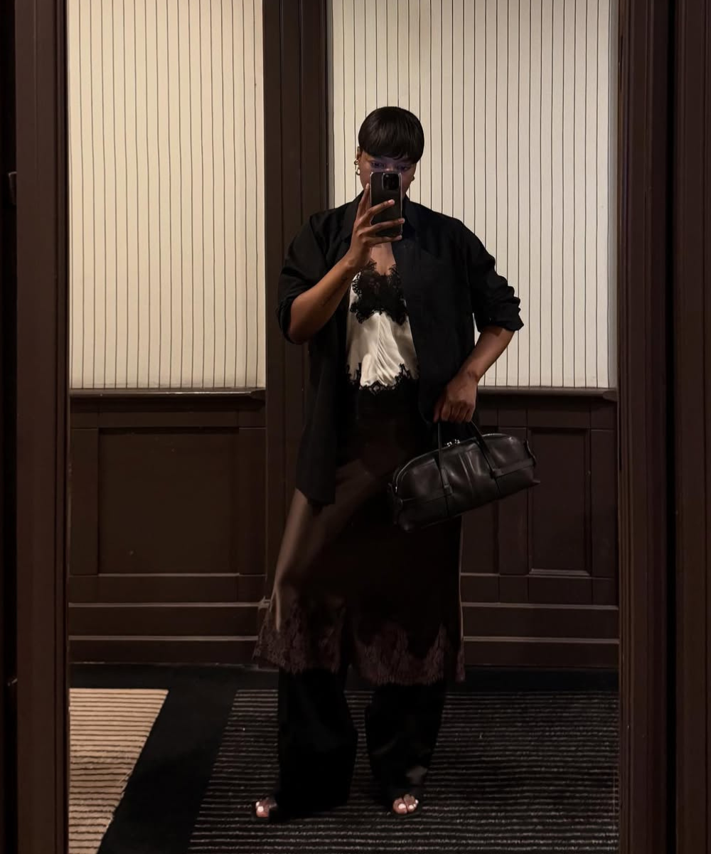 Pessoa tira foto no espelho com celular. Veste blazer preto, segura bolsa e está em local com paredes listradas.