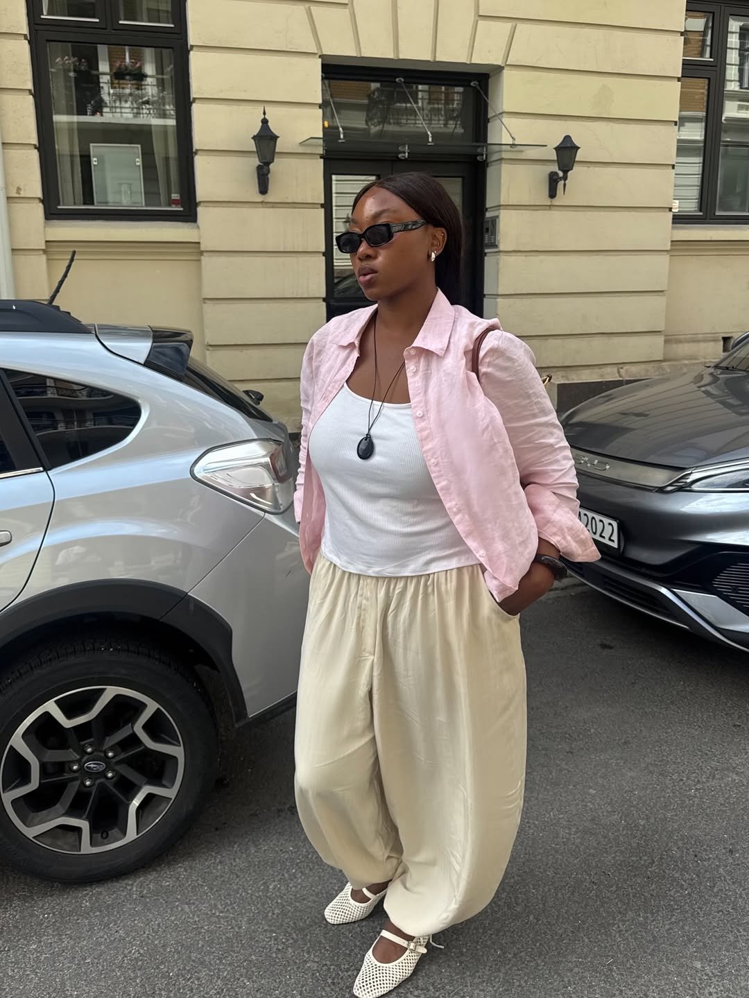 Mulher usa calça balonê bege, blusa branca, camisa rosa e óculos de sol, andando na calçada.