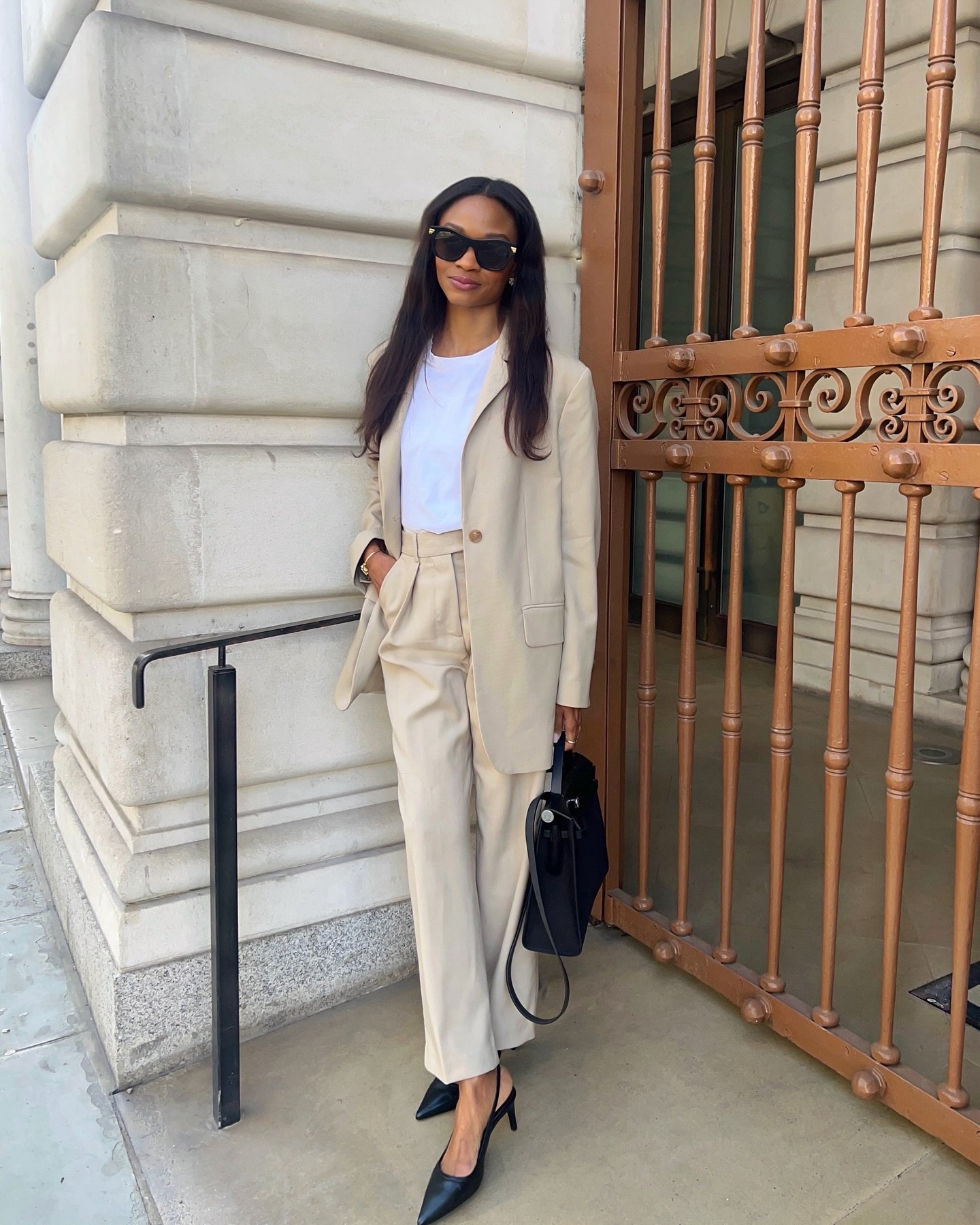 Pessoa com óculos de sol usando blazer e calça social bege, camiseta branca e sapatos pretos, segurando bolsa preta.
