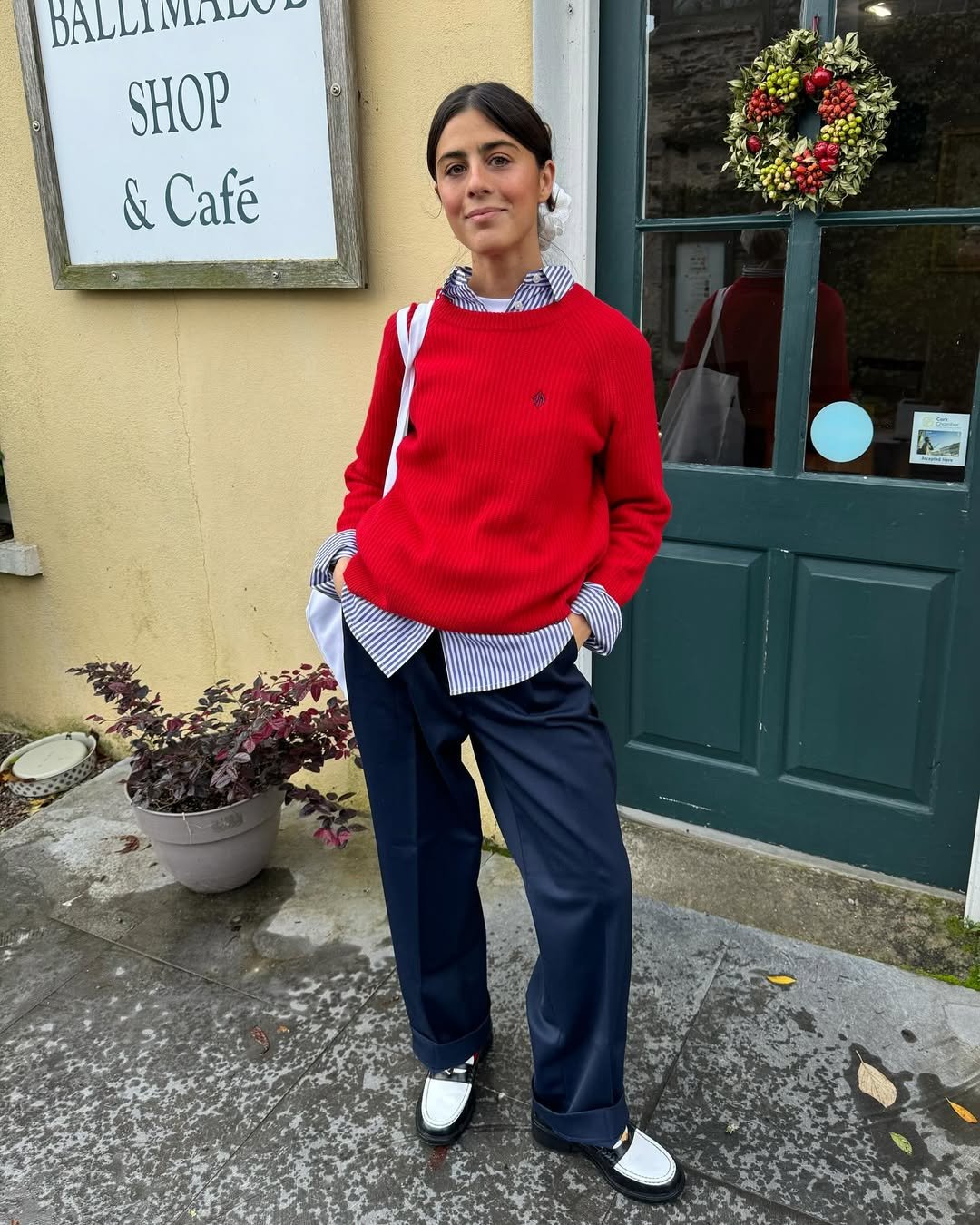 Pessoa usa suéter vermelho, camisa listrada azul e sapato bicolor em frente a uma porta verde com guirlanda.