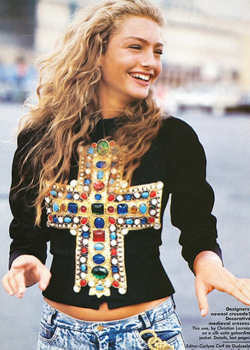 Modelo sorridente com blusa de cruz ornamentada por Christian Lacroix. Cabelo ondulado e jeans.