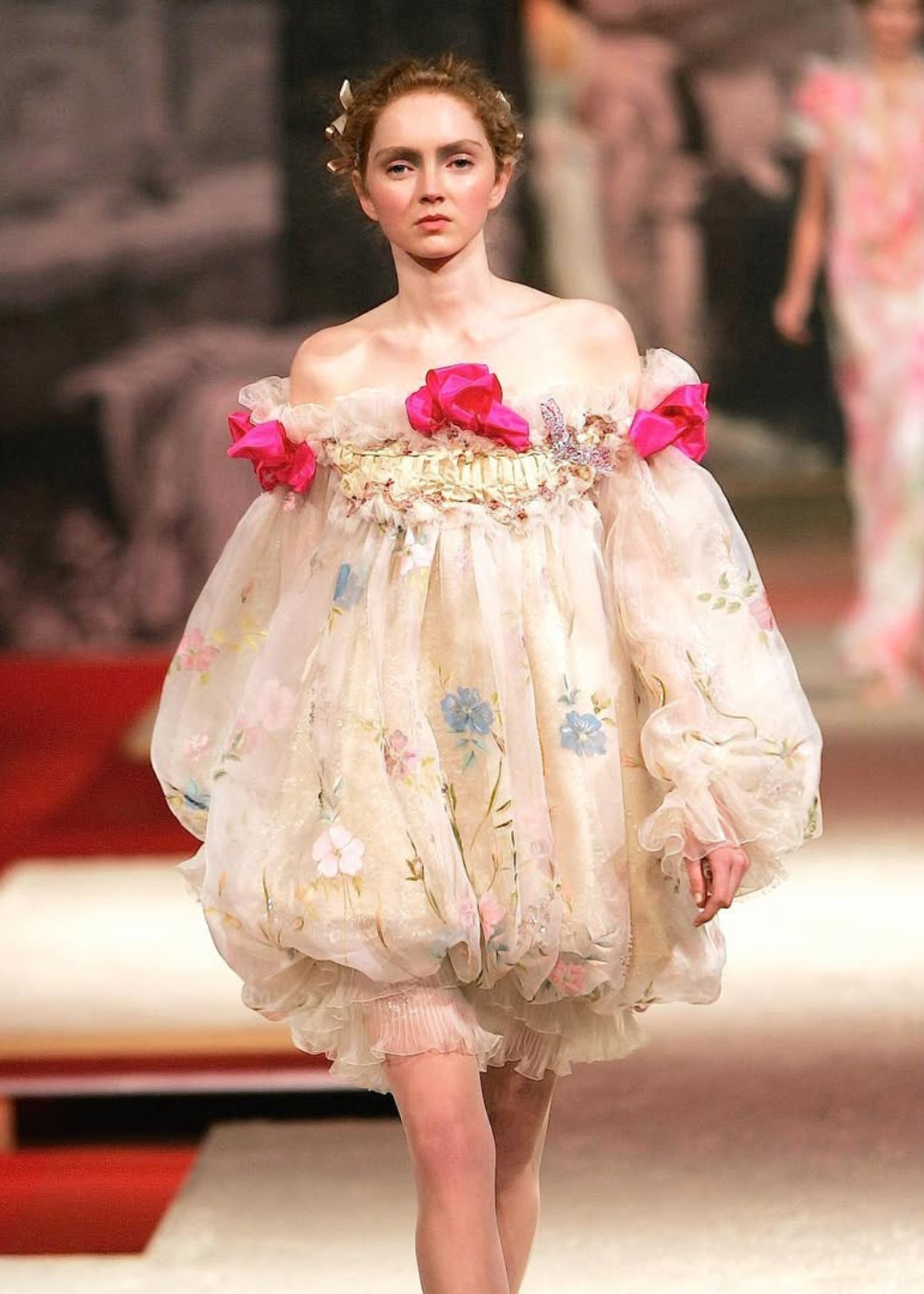 Modelo desfilando em passarela usando vestido floral bufante da coleção de Christian Lacroix, com laços rosas vibrantes.