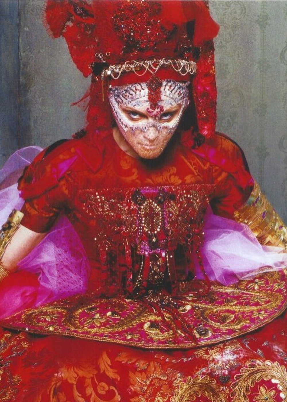 Vestimenta elaborada com máscara e detalhes ricos em vermelho e dourado, evocando estilo teatral e extravagante.