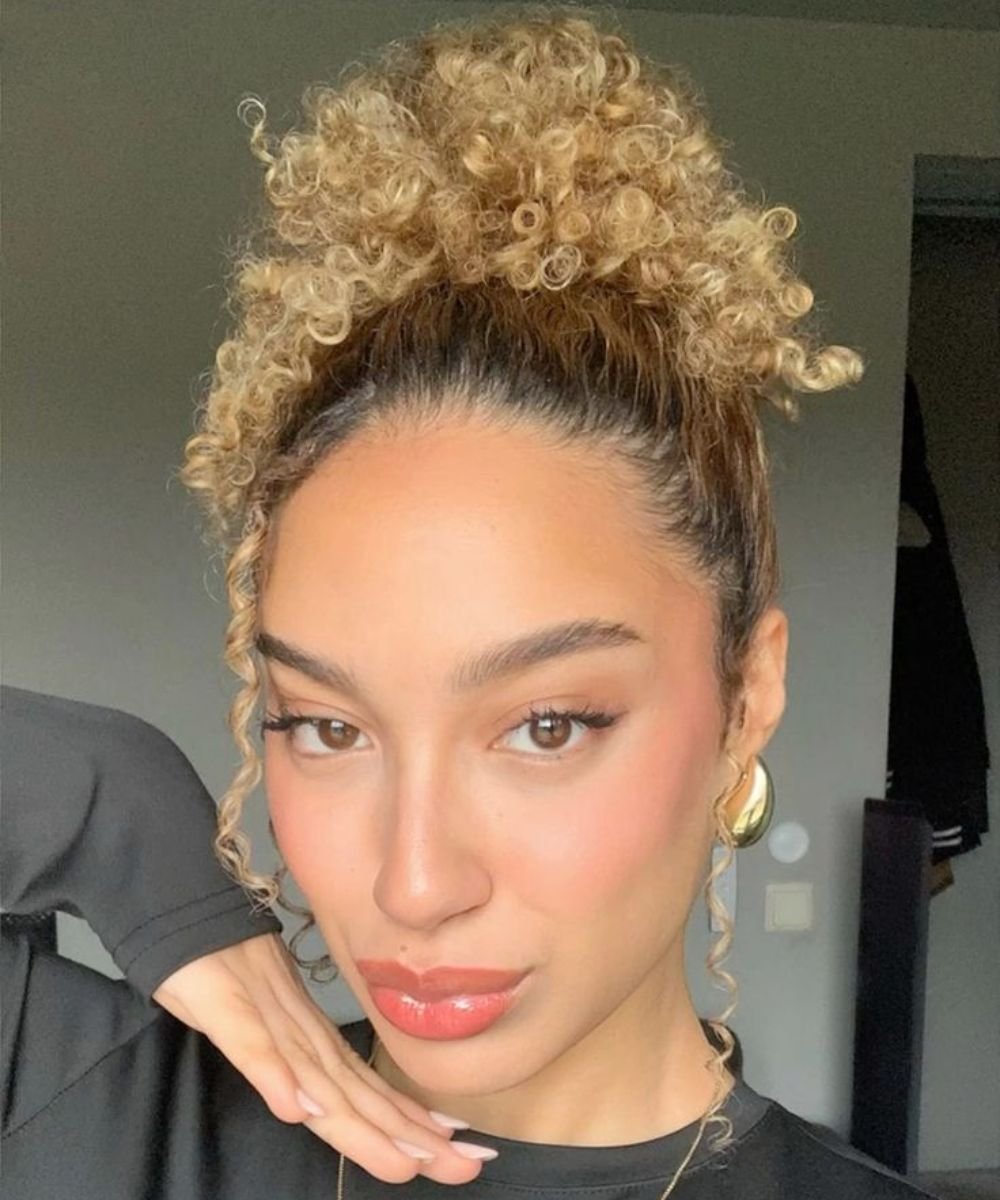 Cabelo cacheado em coque abacaxi, melhor penteado para day after. Maquiagem suave realça os olhos e lábios brilhosos.