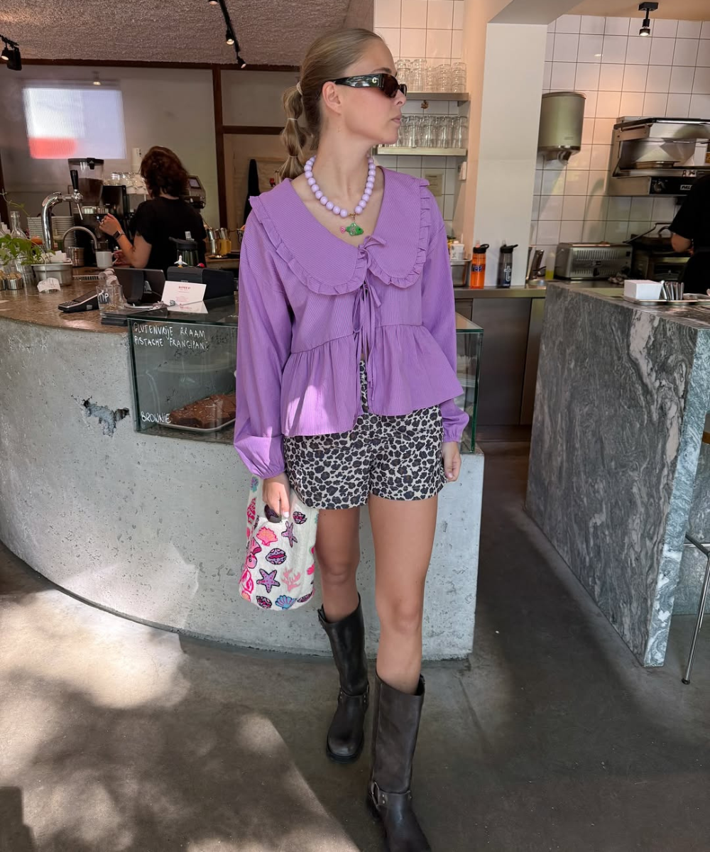 Jovem em café usa blusa roxa e shorts estampados, com bolsa decorada, botas e colar de miçangas.