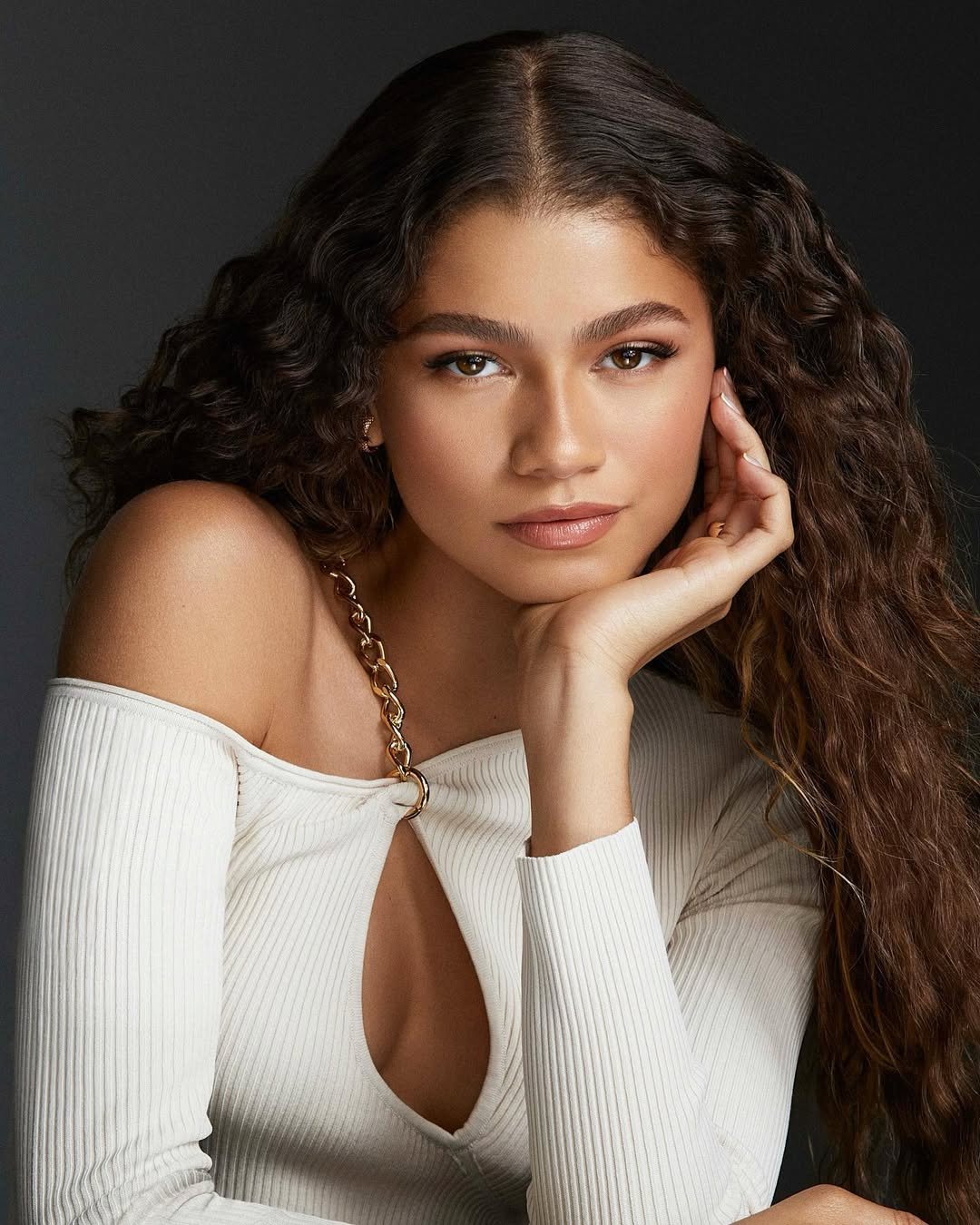 Zendaya de cabelo cacheado apoia o rosto na mão, vestindo blusa branca. Os cabelos brilhantes destacam um bom day after para cabelos cacheados.