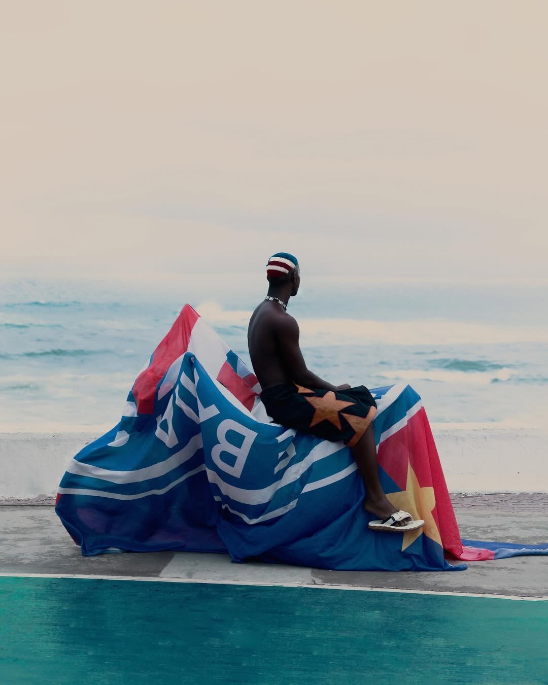 Pessoa sentada à beira-mar, vestindo tons patrióticos e envolta em bandeira colorida.Dendezeiro