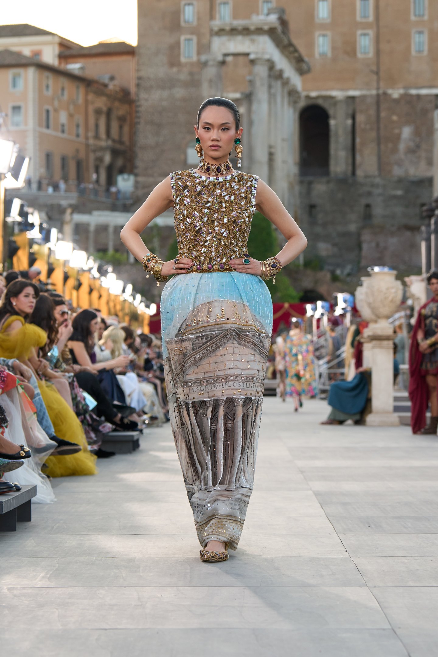 Modelo usa vestido Dolce&Gabanna dourado e azul com detalhes arquitetônicos, desfilando em ambiente histórico ao ar livre.