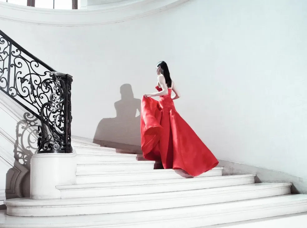 Mulher em vestido vermelho sobe elegante escadaria branca com corrimão ornamental preto. Elegância e moda. Dior e Eu