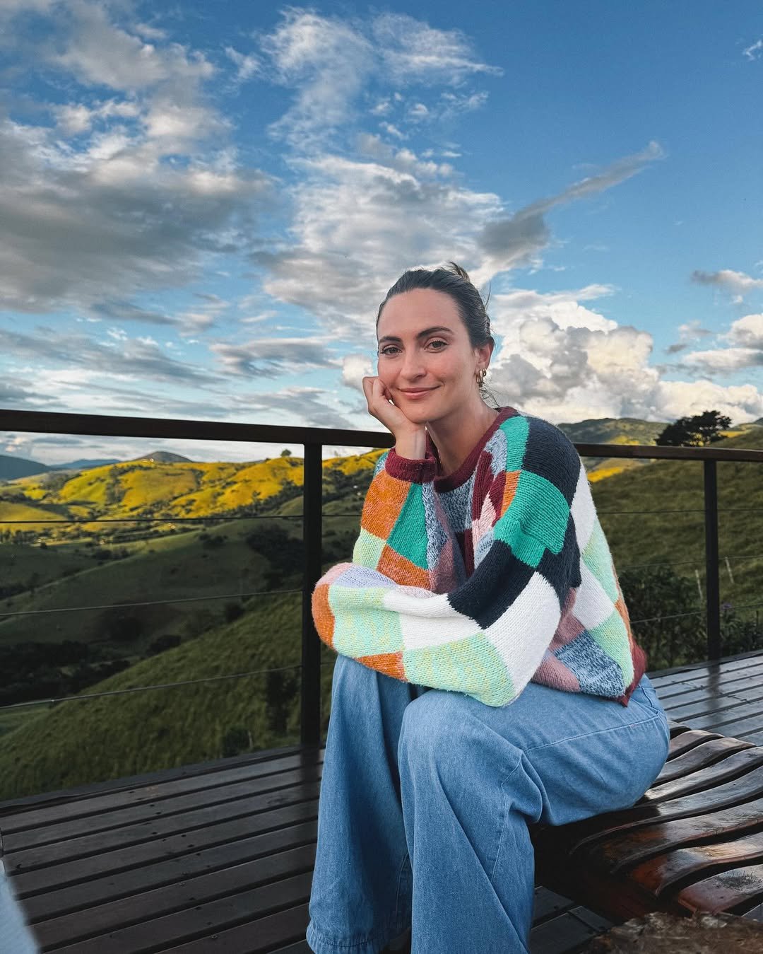 Pessoa sorrindo em deck de madeira com vista para colinas verdejantes sob céu azul e nublado. Como organizar a mente