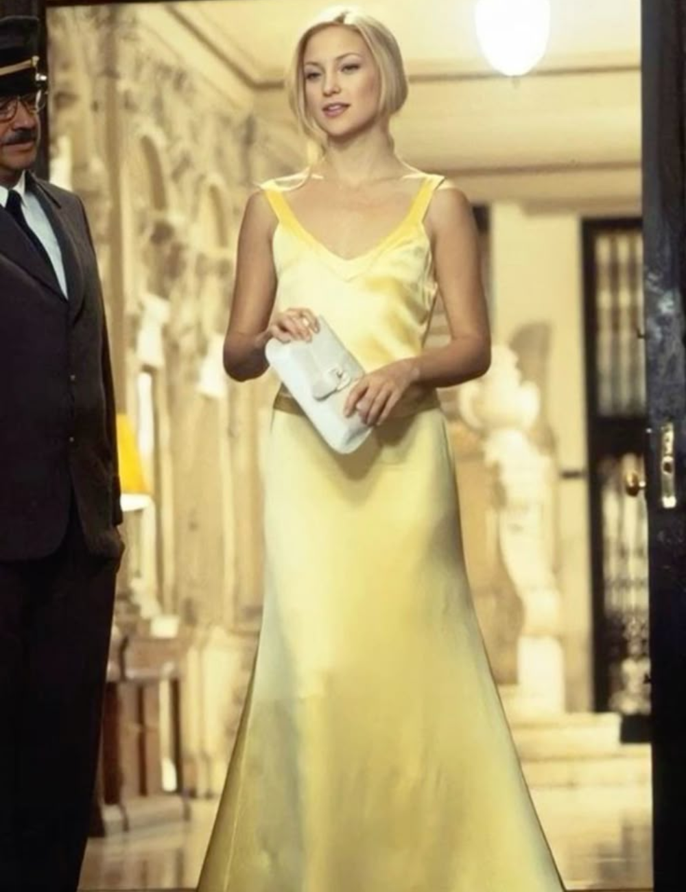 Pessoa usando vestido amarelo elegante e segurando bolsa, em cenário lembrando comédias românticas.