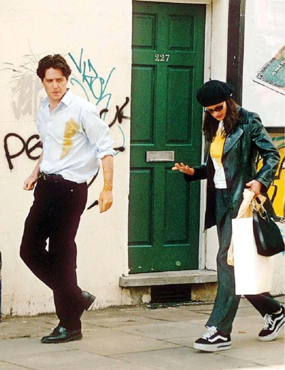 Homem com mancha na camisa e mulher com chapéu andam em rua grafitada, evocando cenas de comédias românticas.