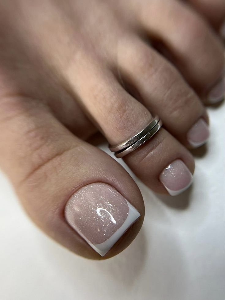 Unhas do pé com esmalte estilo francesinha no pé em tons claros e anel prateado no dedo do pé.