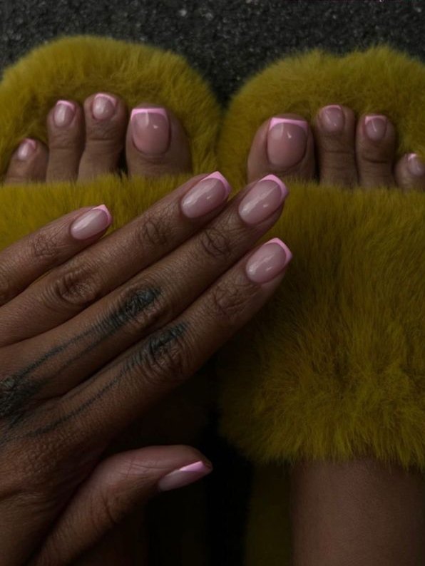 Unhas com esmalte rosa e francesinha no pé são mostradas, usando chinelos peludos amarelos.