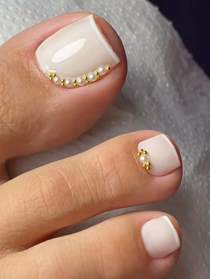 Francesinha no pé com detalhes de pérolas e apliques dourados nas unhas, sobre tecido branco.