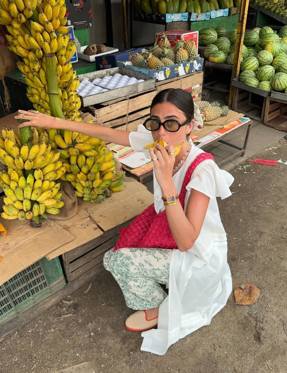 Mulher comendo banana ajoelhada em frente a barraca de frutas, usando vestido branco e friulanes bege.