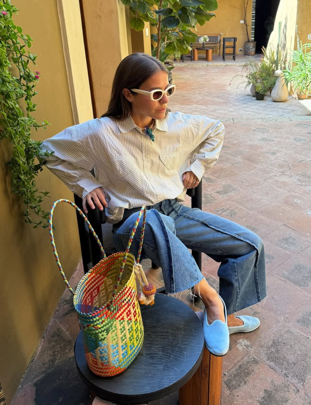 Pessoa com óculos escuros sentada ao lado de uma sacola colorida, vestindo 