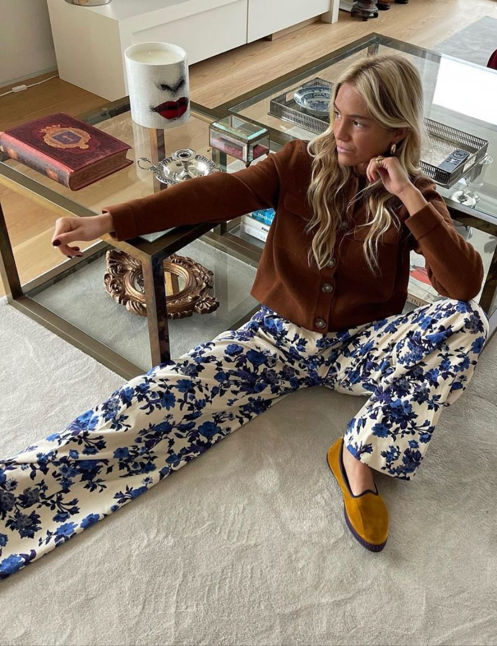 Mulher sentada em sala, vestindo camisa marrom e calça floral azul; calça Friulanes mostarda.