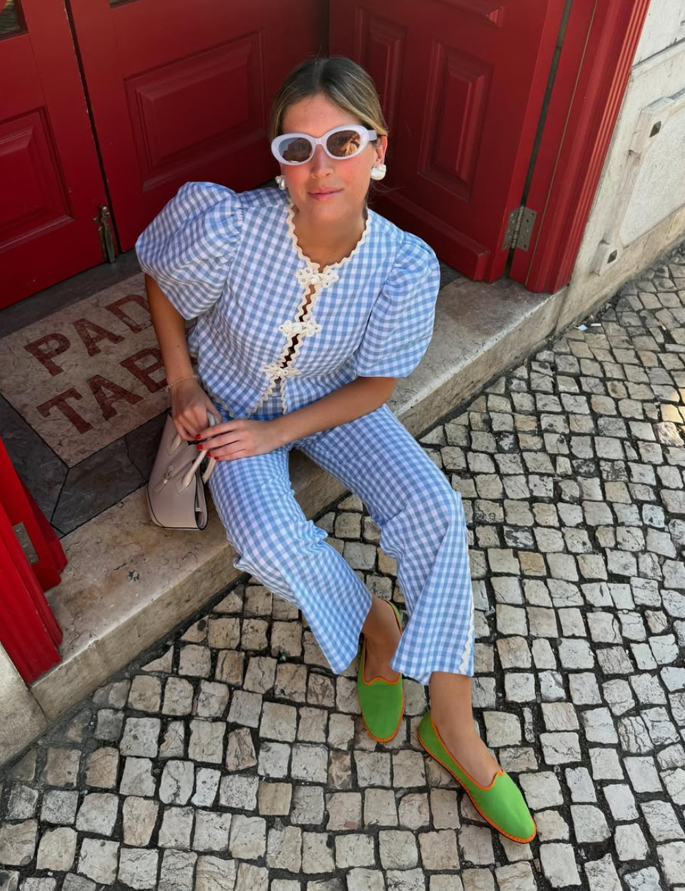 Pessoa sentada em calçada de pedras usando óculos de sol, roupa xadrez azul e sapatos verdes estilo friulanes.