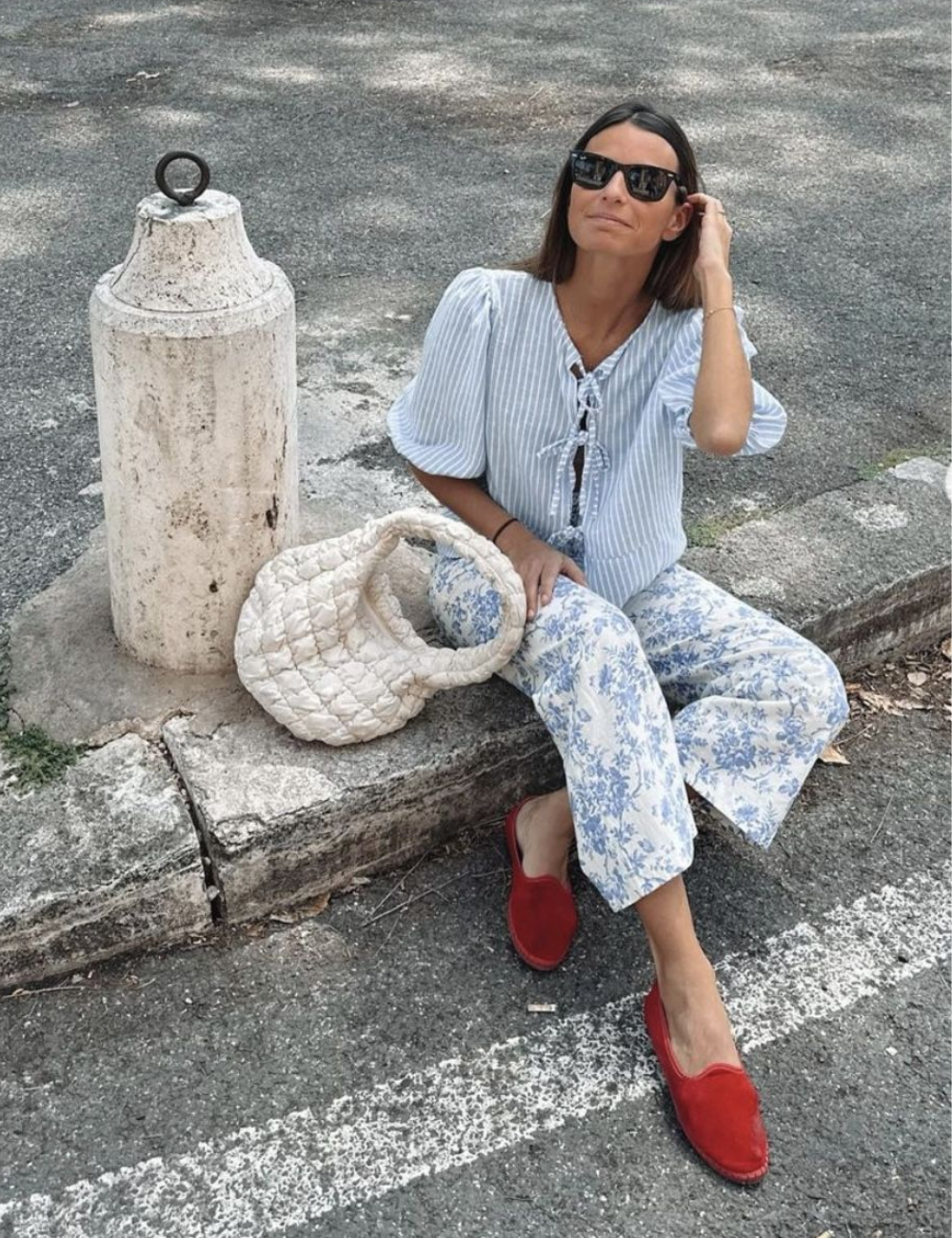 Mulher sentada com calças florais e sapatos vermelhos friulanes, bolsa branca ao lado, usa óculos escuros.