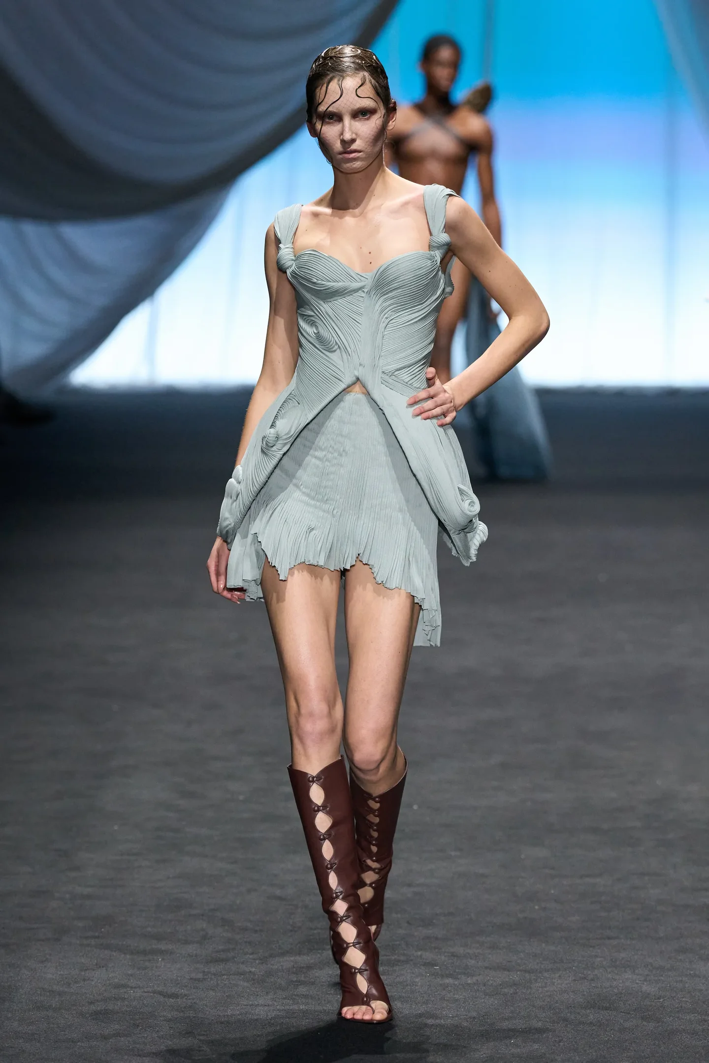 Modelo desfila com vestido cinza claro e sandálias gladiadora em uma passarela.